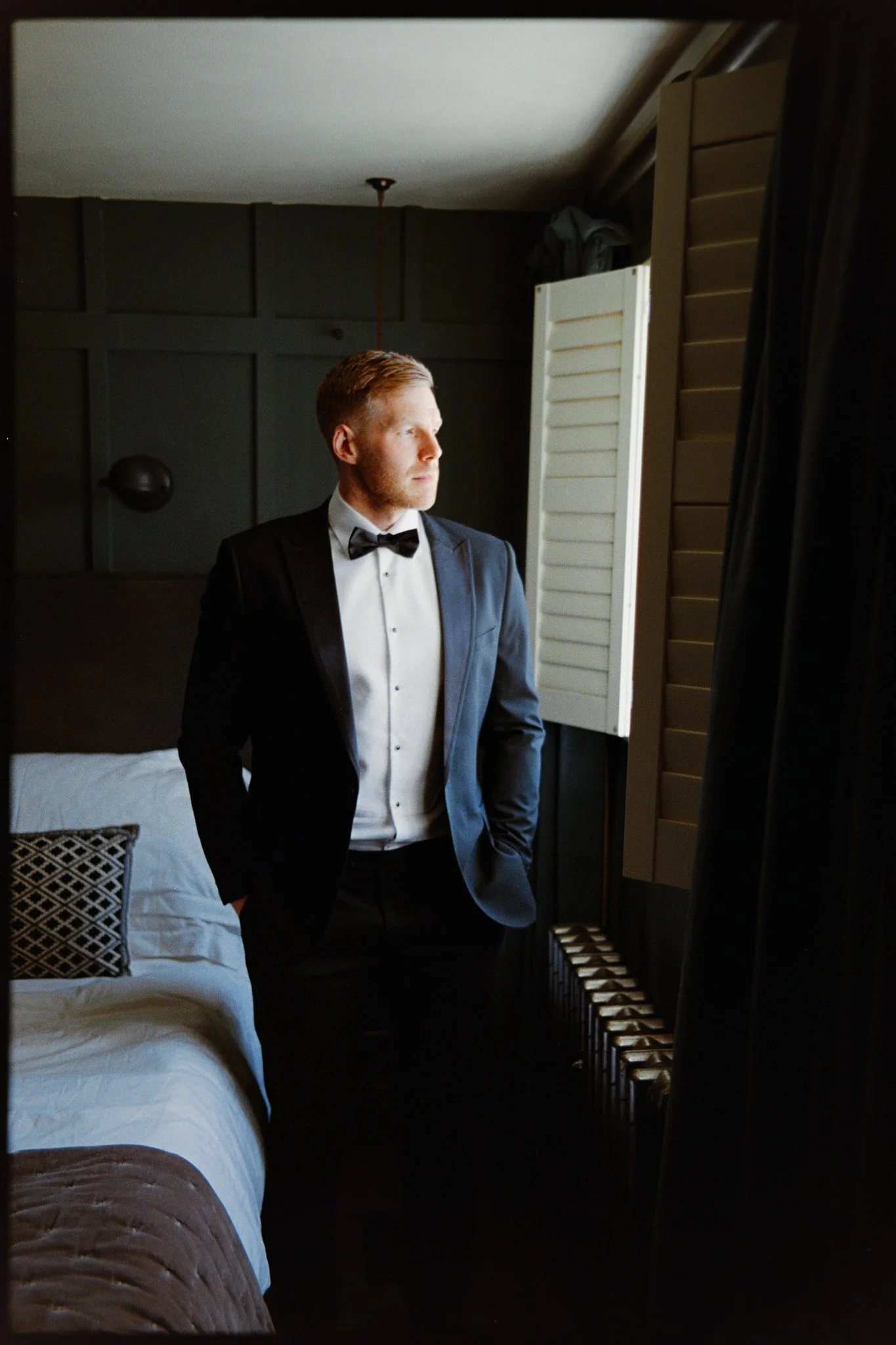 A man in formal attire, wearing a tuxedo and bow tie, standing in a hotel room near a window with shutters, looking out with a serious expression.