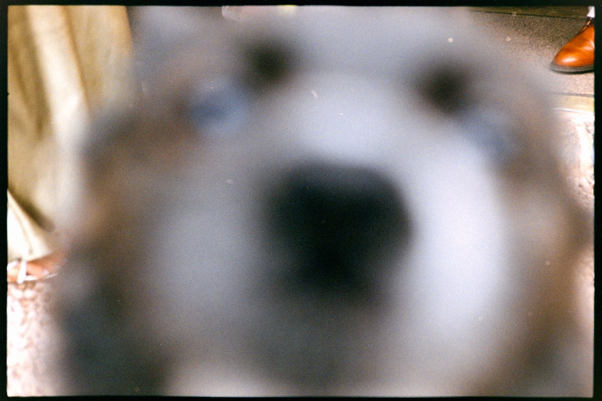 Close-up of a black device or object with blurry black spots, with part of a brown shoe visible on the top right corner, and a beige surface on the left side.