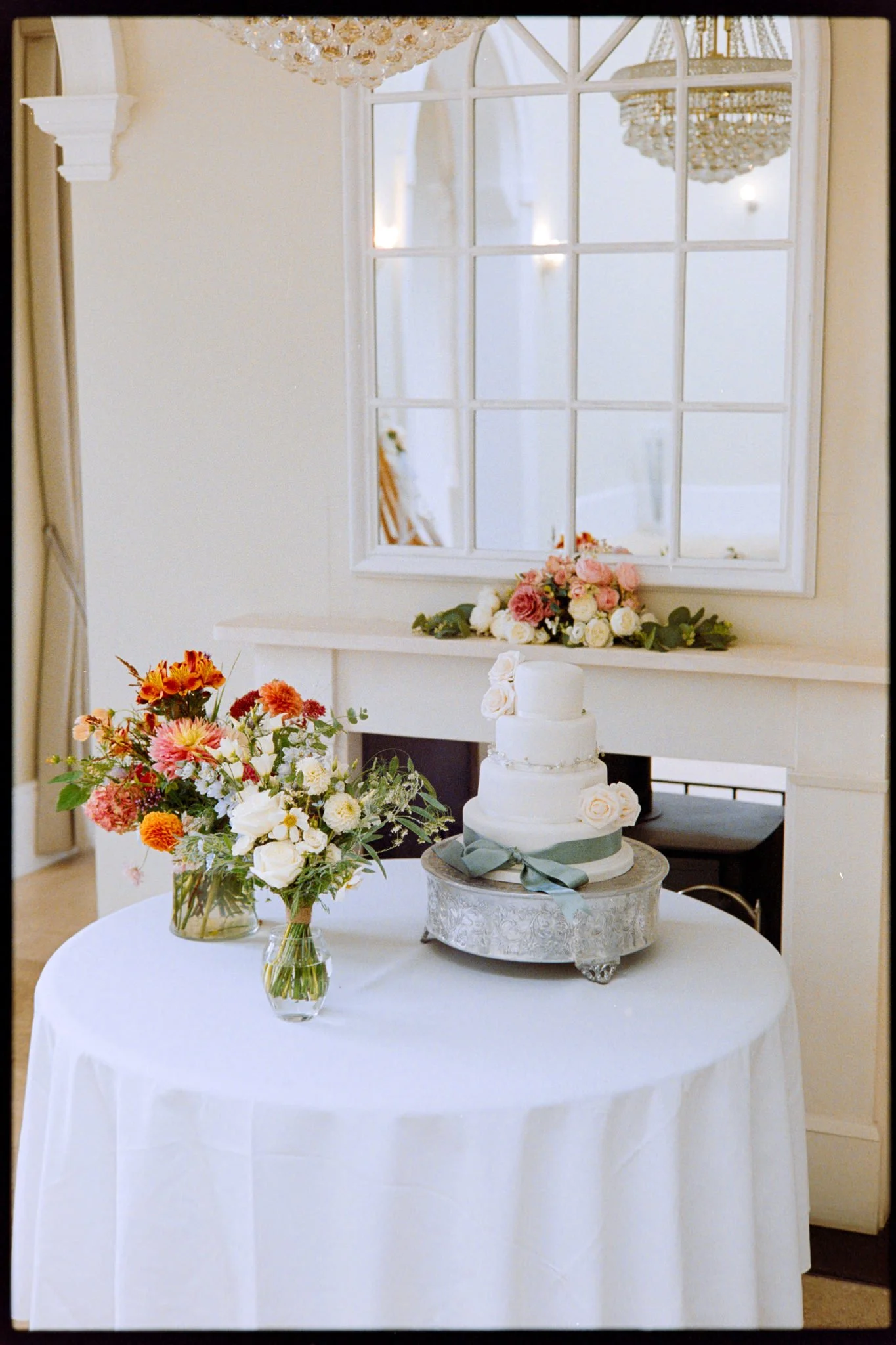A white round table with a floral arrangement in a glass vase, a white multi-tiered cake with gray ribbon and roses, set in a room with a large window and chandeliers, decorated for a celebration.