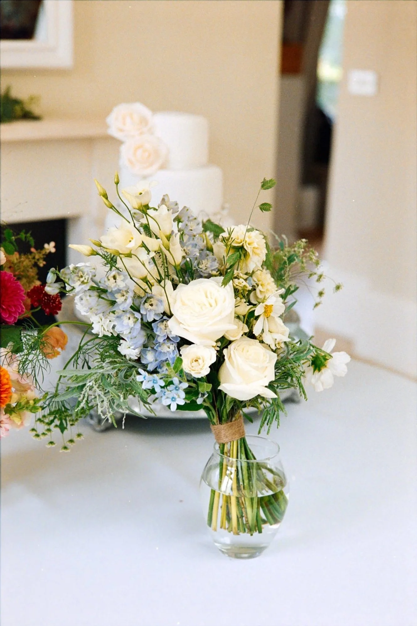 A bouquet of white and pale yellow flowers in a clear glass vase on a white surface, with a blurred background of an indoor setting.