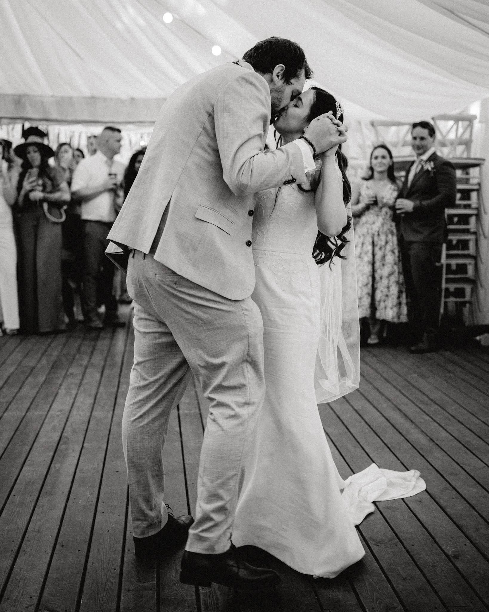 A couple shares a kiss on their wedding dance floor, surrounded by friends and family at a wedding reception.