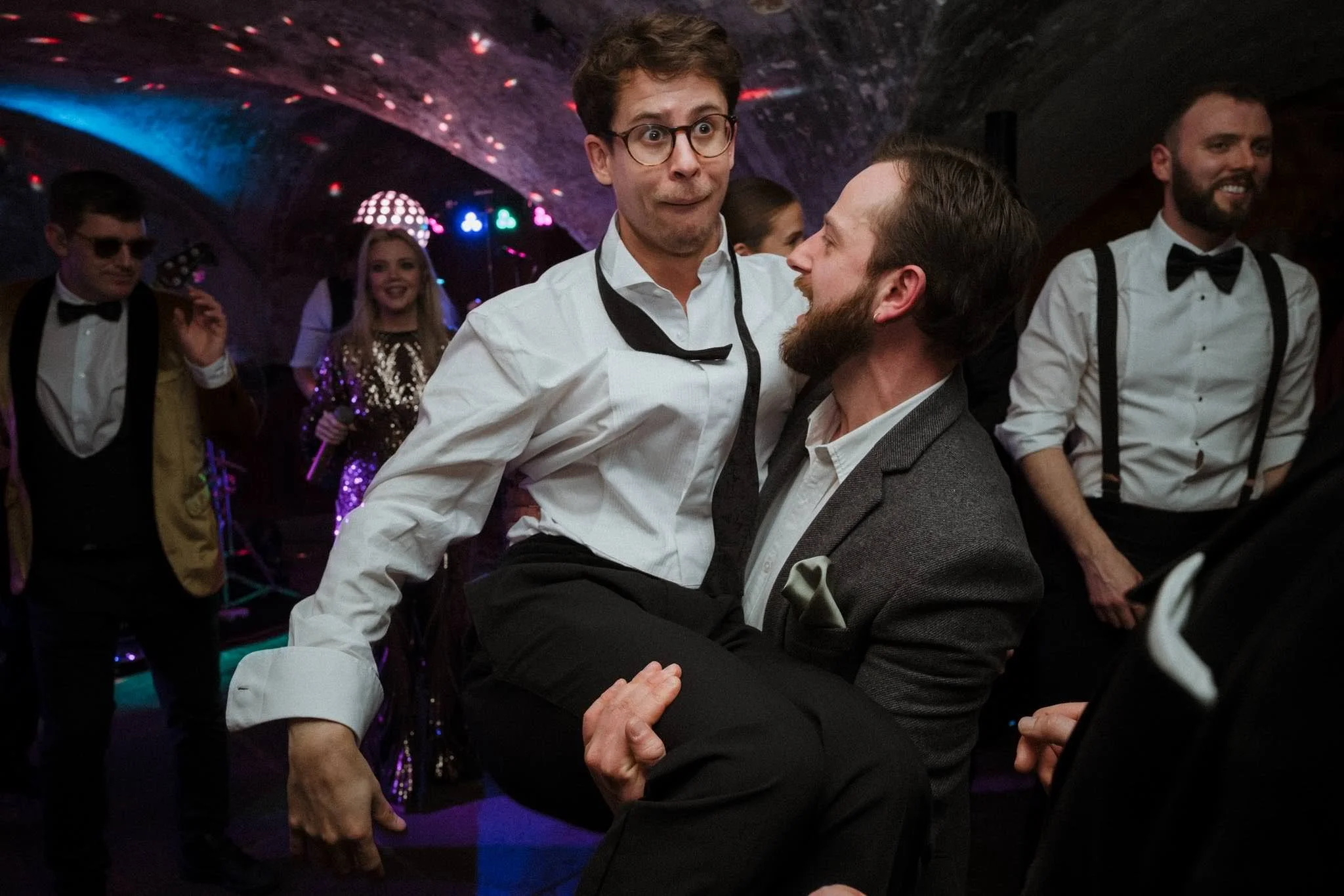 Two men dancing closely together at a party or dance event, with other people in formal attire in the background.