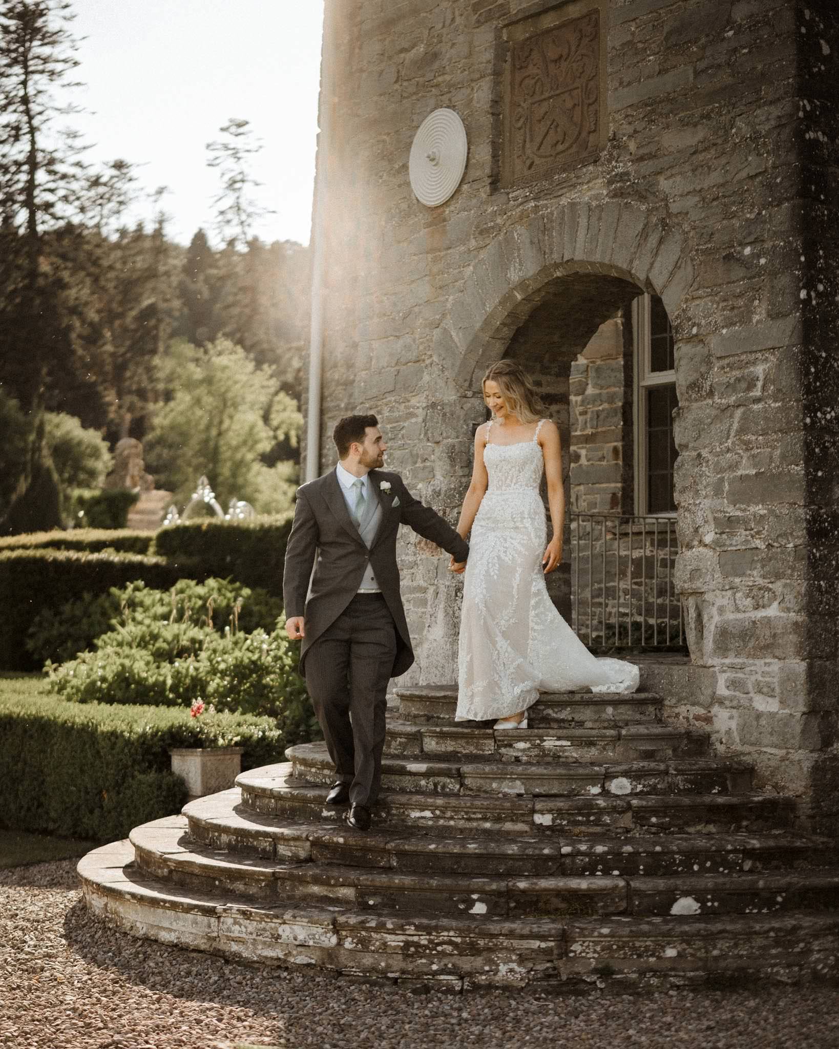 A bride and groom holding hands and smiling as they walk down the steps of a stone building. The bride is wearing a white wedding dress, and the groom is in a gray suit. The scene is outdoors with greenery and sunlight.