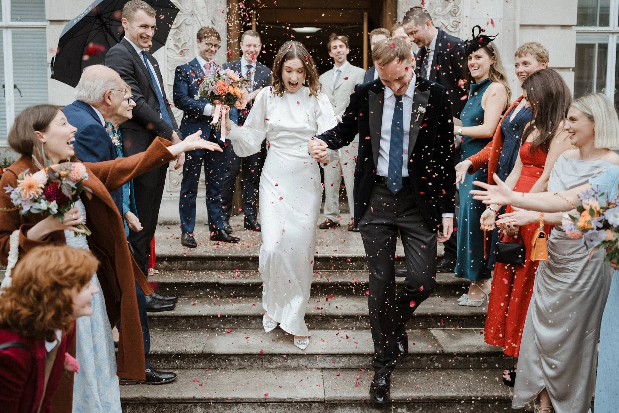 A bride and groom walking down the steps of a building, holding hands, surrounded by friends and family throwing confetti, celebrating their wedding.