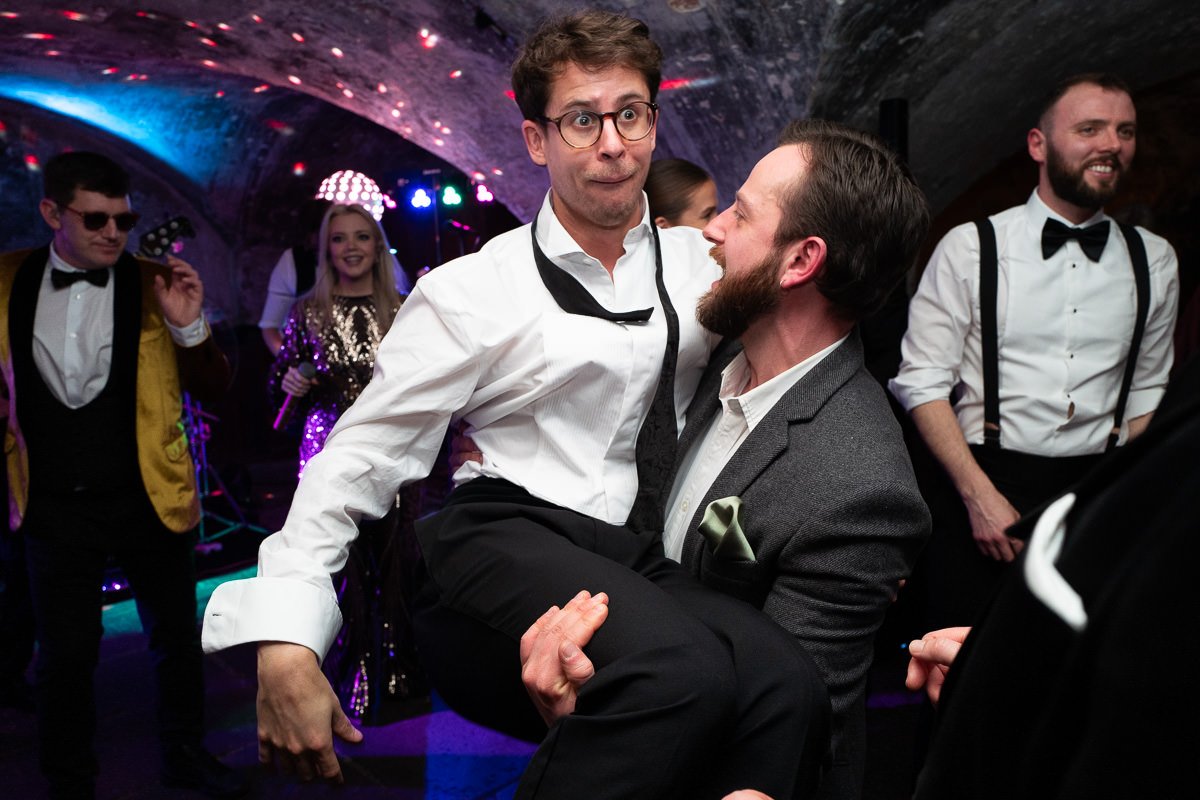 Groomsman hoisted by friends on the dancefloor at The Mount Without Crypts, pulling a hilarious face mid-air — candid colour wedding reportage