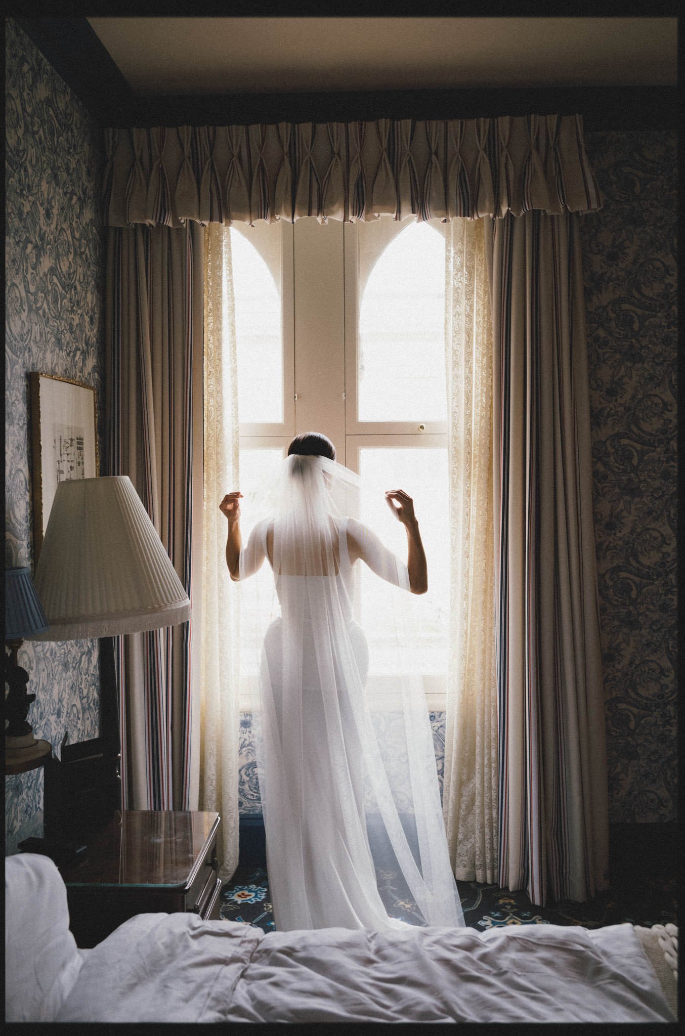 A bride poses by a large georgian window and adjusts her veil at the Randolph Hotel in OXford
