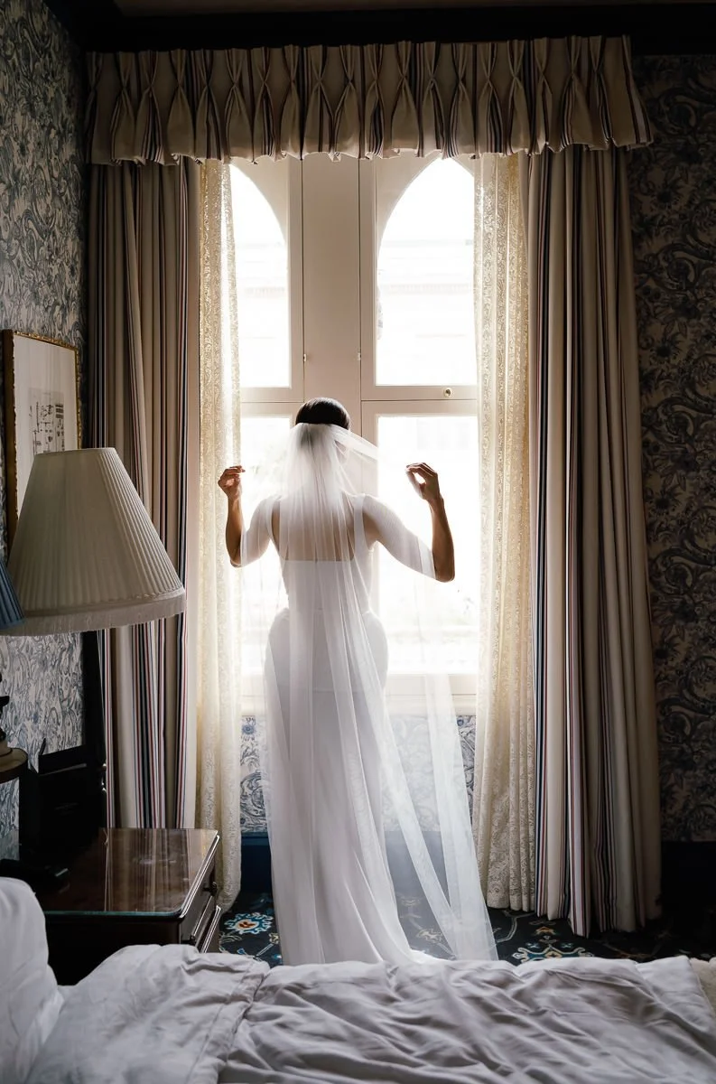 Bride adjusts her veil while gazing out of a window at the Randolph Hotel, Oxford — an intimate colour getting-ready portrait