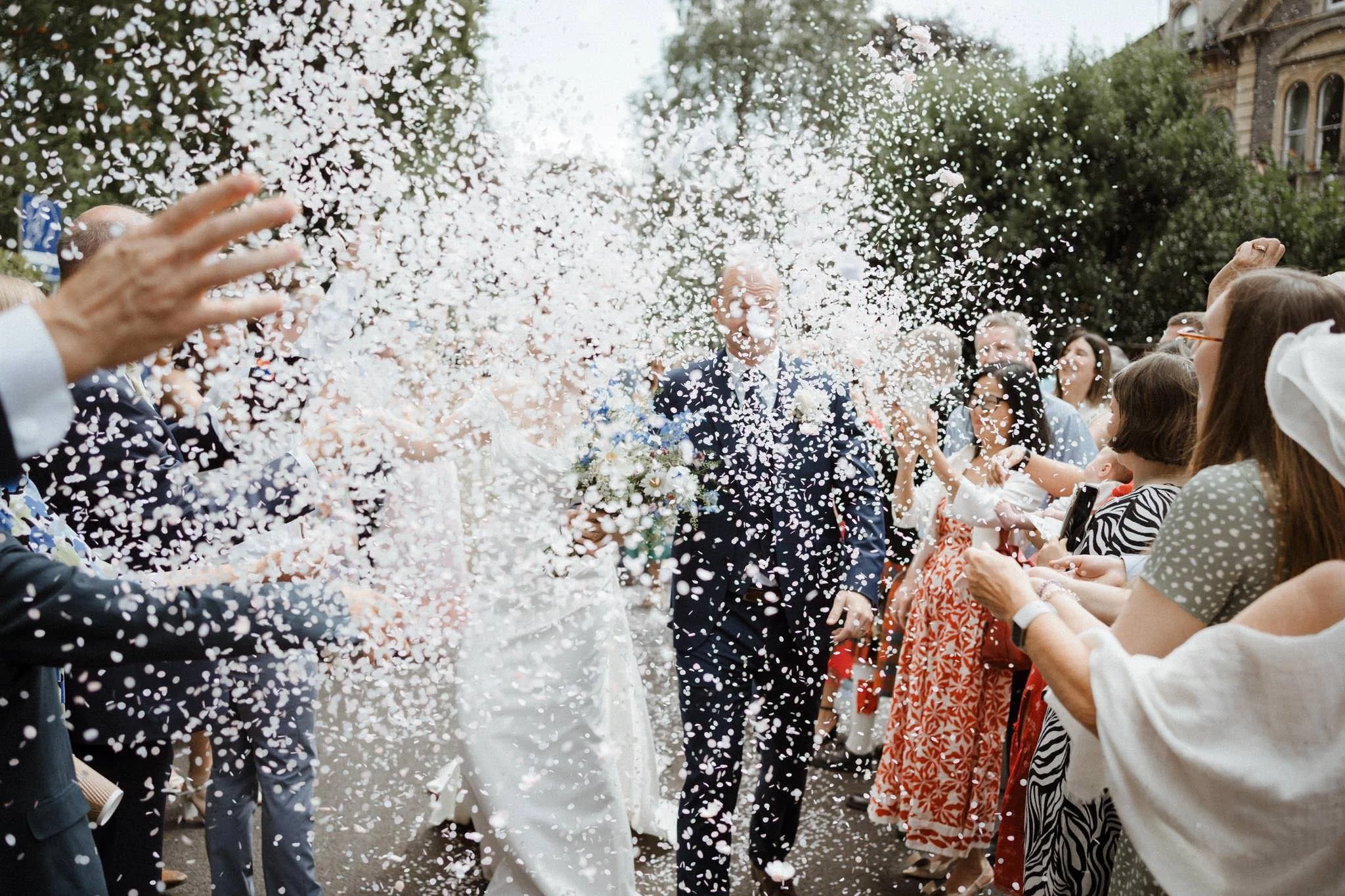 People celebrating with confetti at a wedding, with a bride and groom walking through a crowd of friends and family.