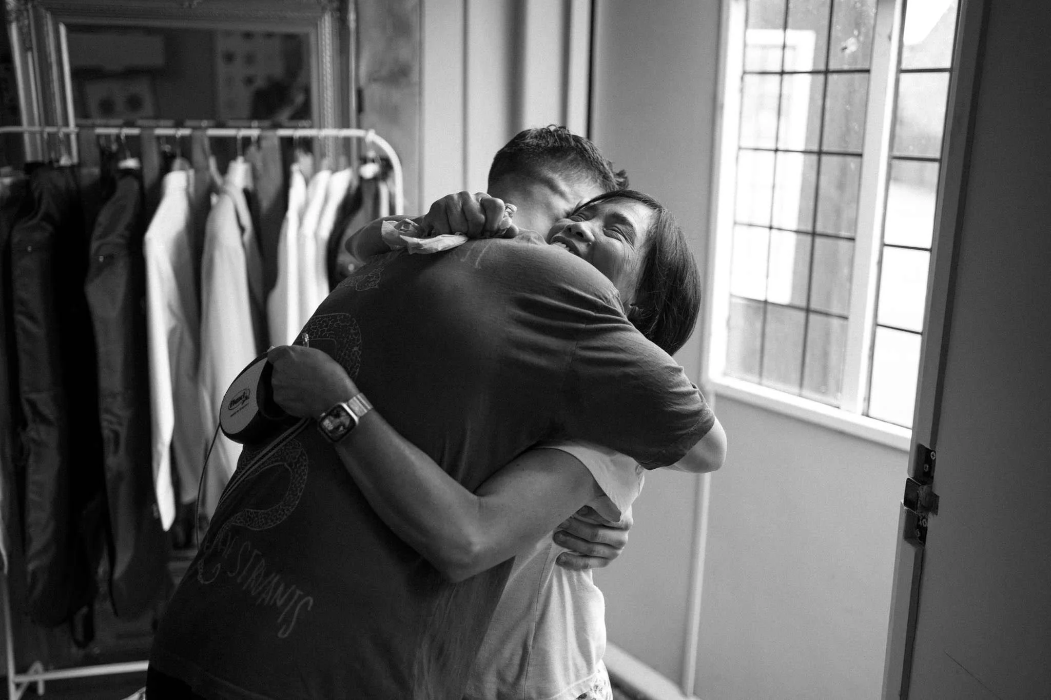 Two people hugging and smiling in a room with clothes hanging on a rack and a window in the background.