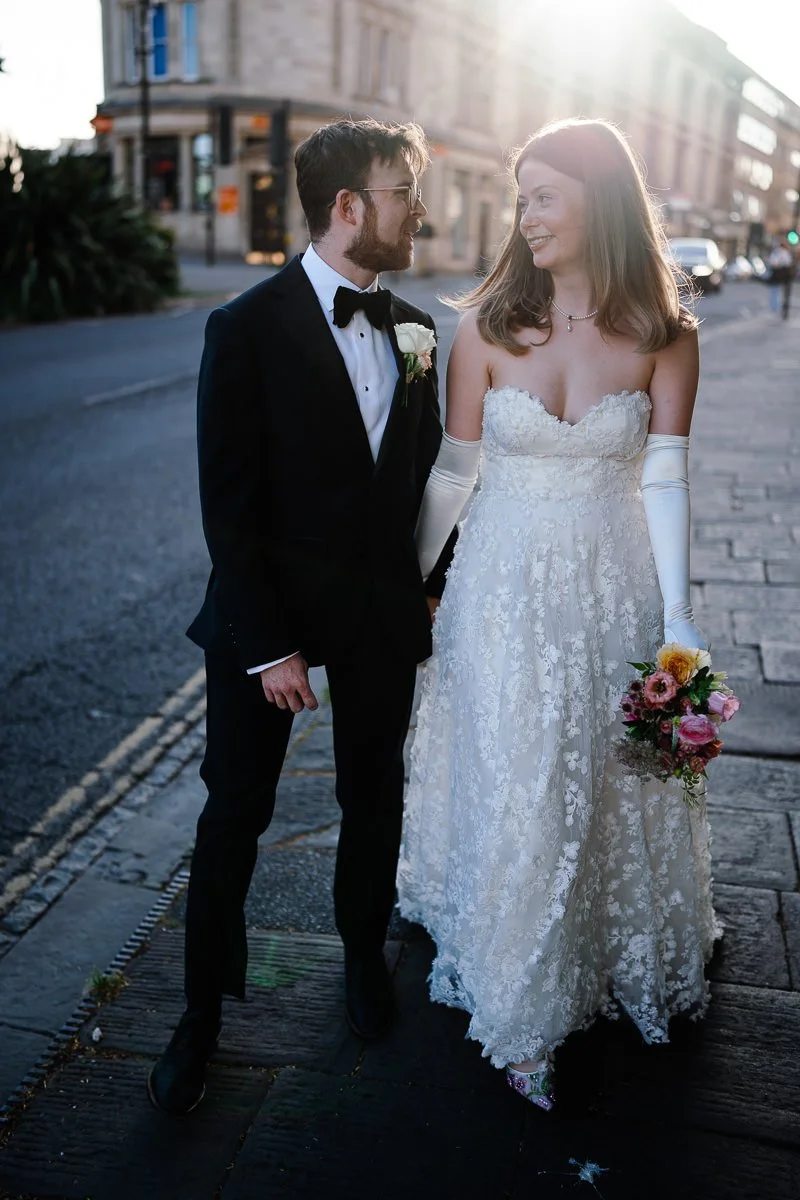 Bride and groom bathed in golden sunlight on the Triangle in Bristol, he in black tie and she in a vintage-style white dress with sleeve gloves