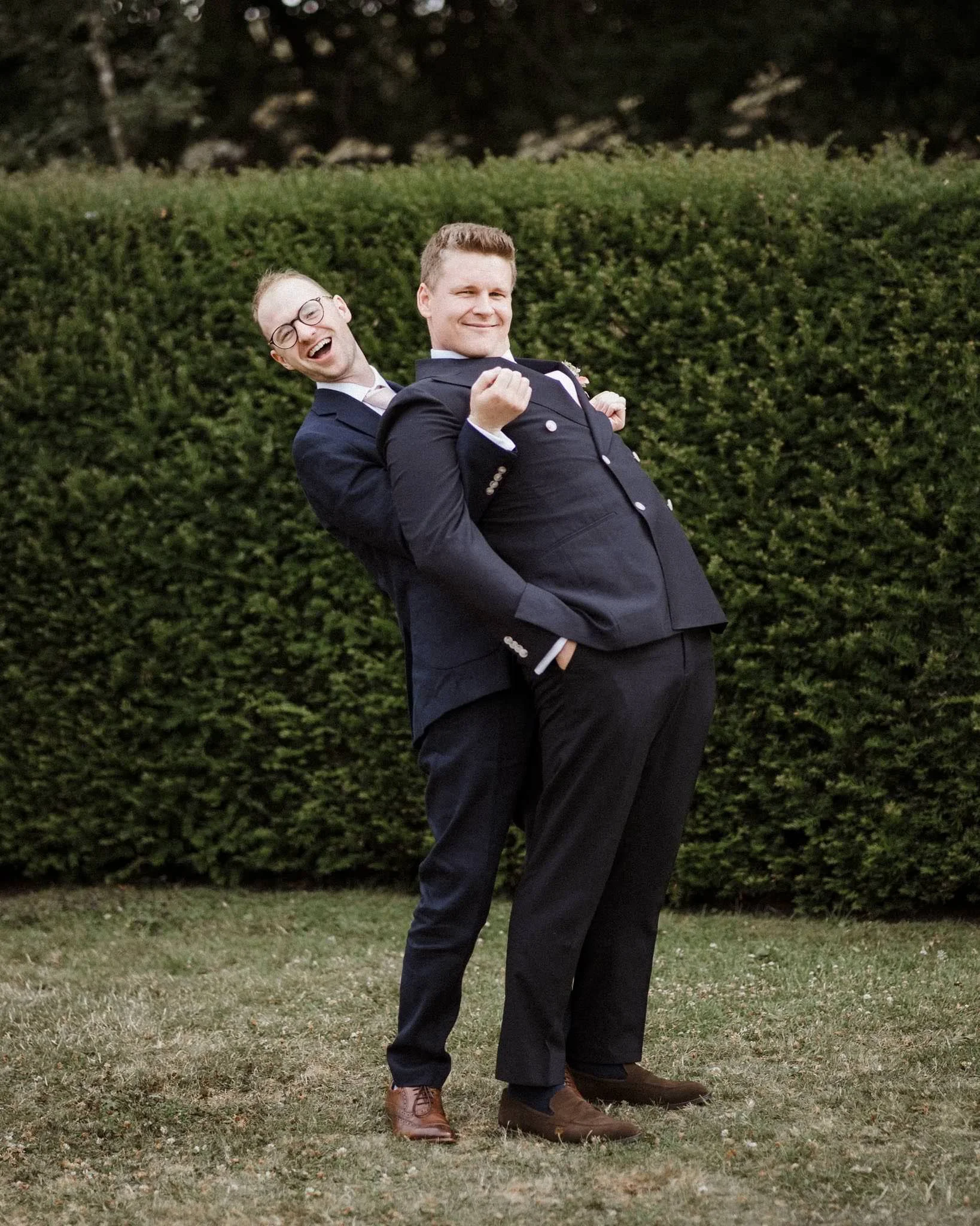 Two men in suits smiling and playfully leaning back against each other outdoors in front of a green hedge.