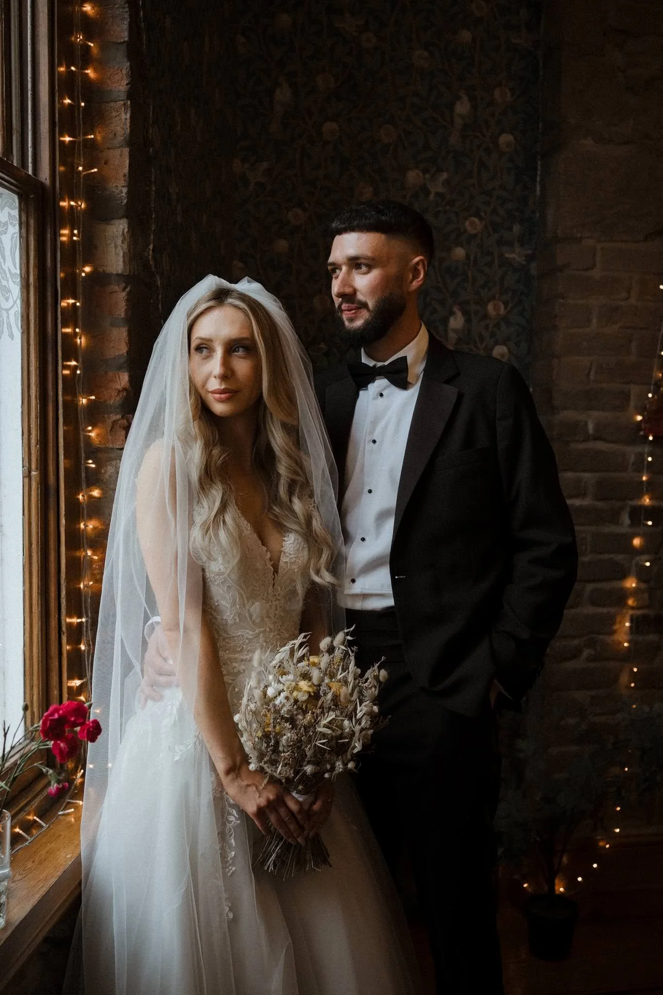Editorial wedding photography of a bride and groom in Bristol standing by a window with black tie and blonde bride and fairy lights behind