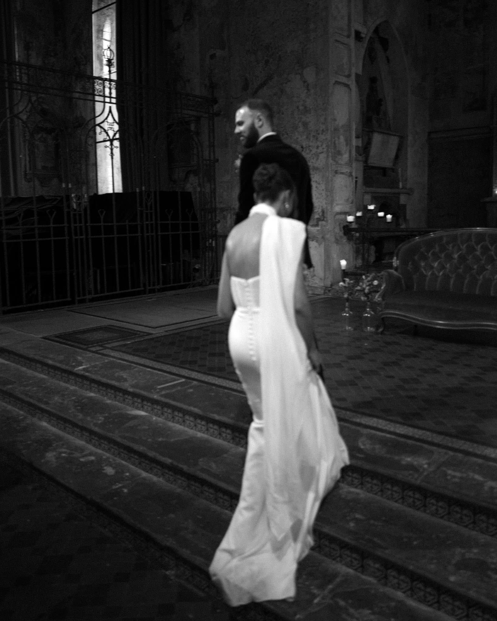 A black and white photo of a man and woman inside a historic, ornate building. The woman is wearing a long, white strapless gown, and the man is dressed in a tuxedo. Candles are placed on a table and a sofa is visible in the background.