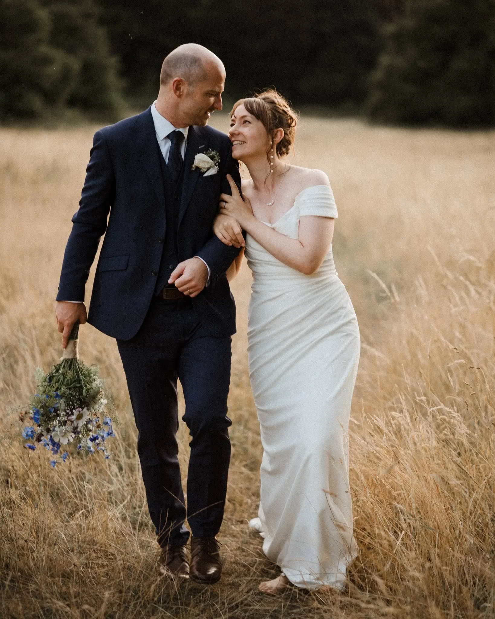 A bride and groom walking through a field of tall golden grass, smiling and looking at each other, with the groom holding a bouquet of flowers.