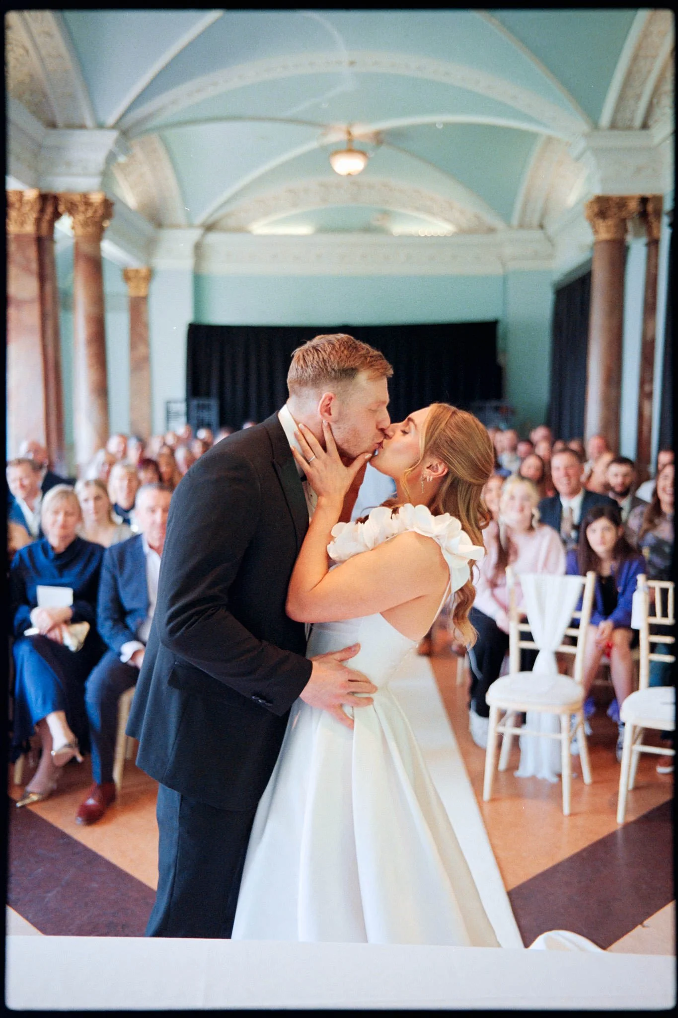 At a wedding ceremony at Cheltenham Town hall, the bride and groom kiss on Kodak film with the guests behind them