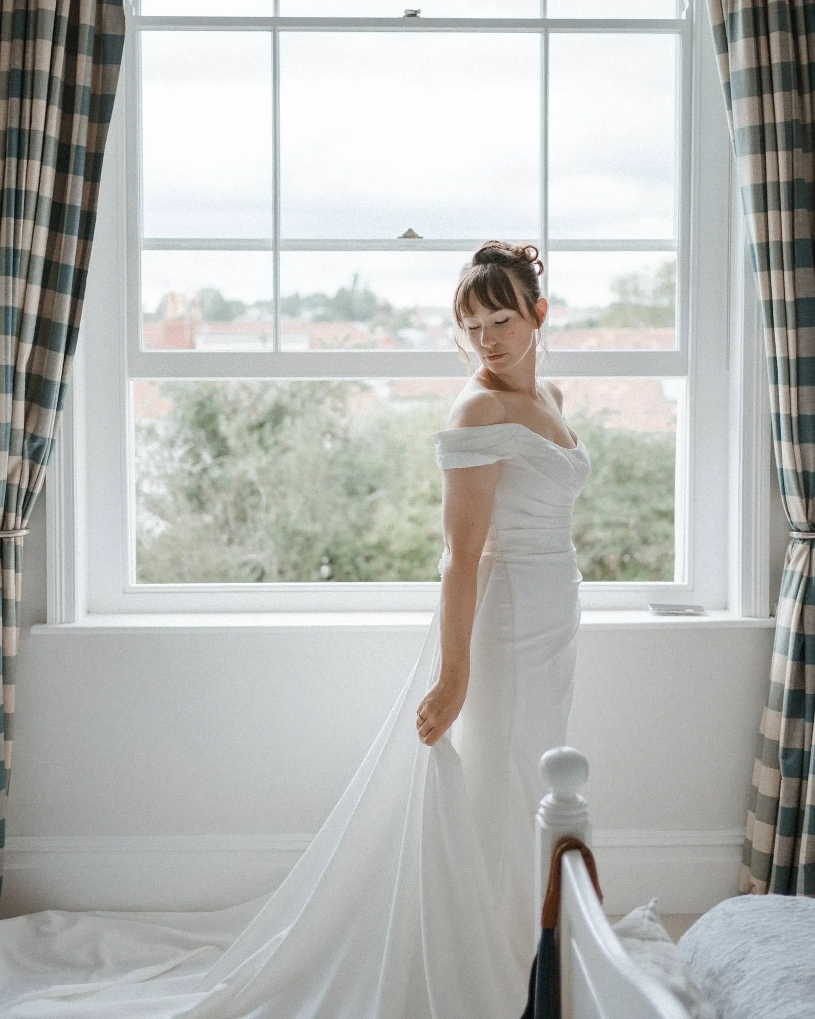 A woman in a white wedding dress standing in front of a large window with curtains, looking down with her eyes closed.