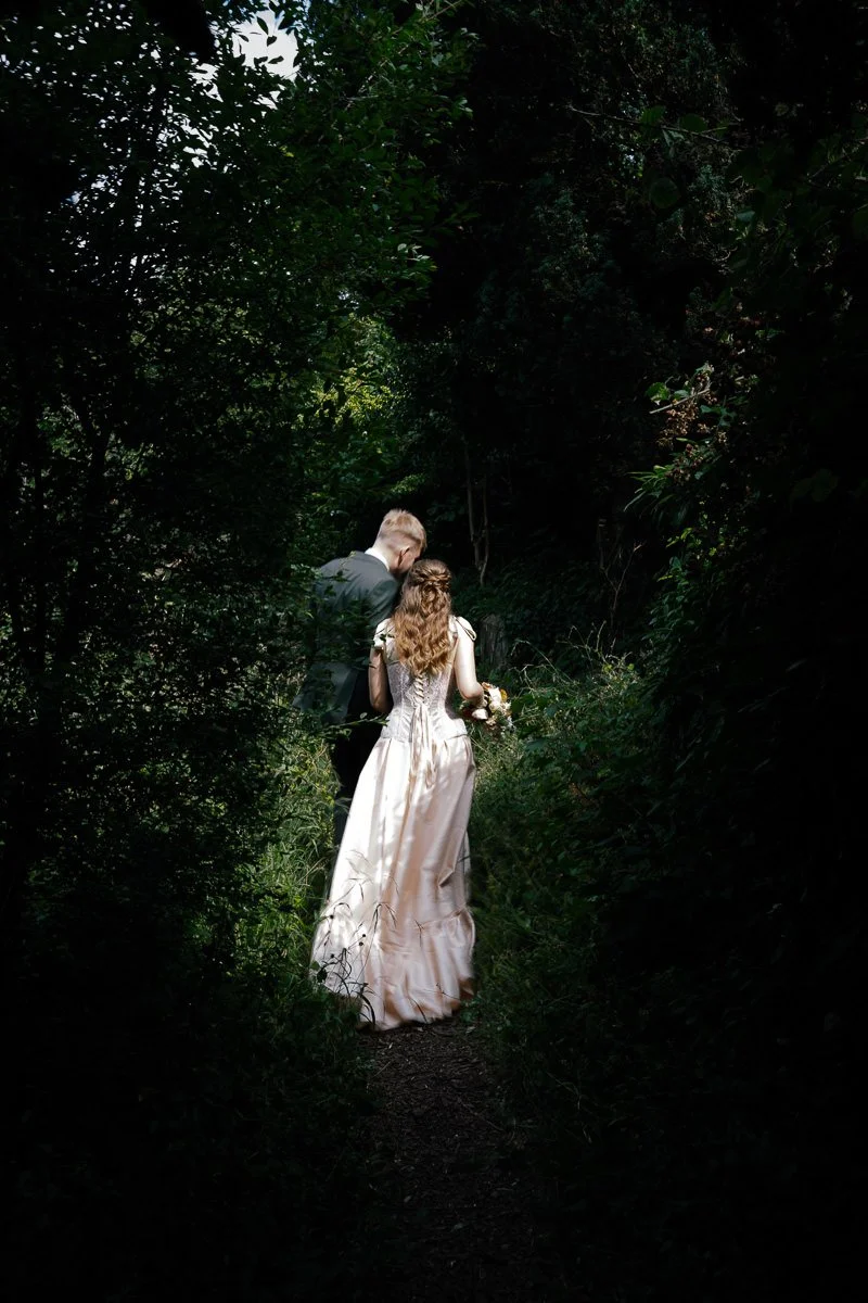 Bride and groom walk into a patch of light at Arnos Vale Cemetery, she in an off-white vintage tie-up dress — a cinematic colour wedding portrait