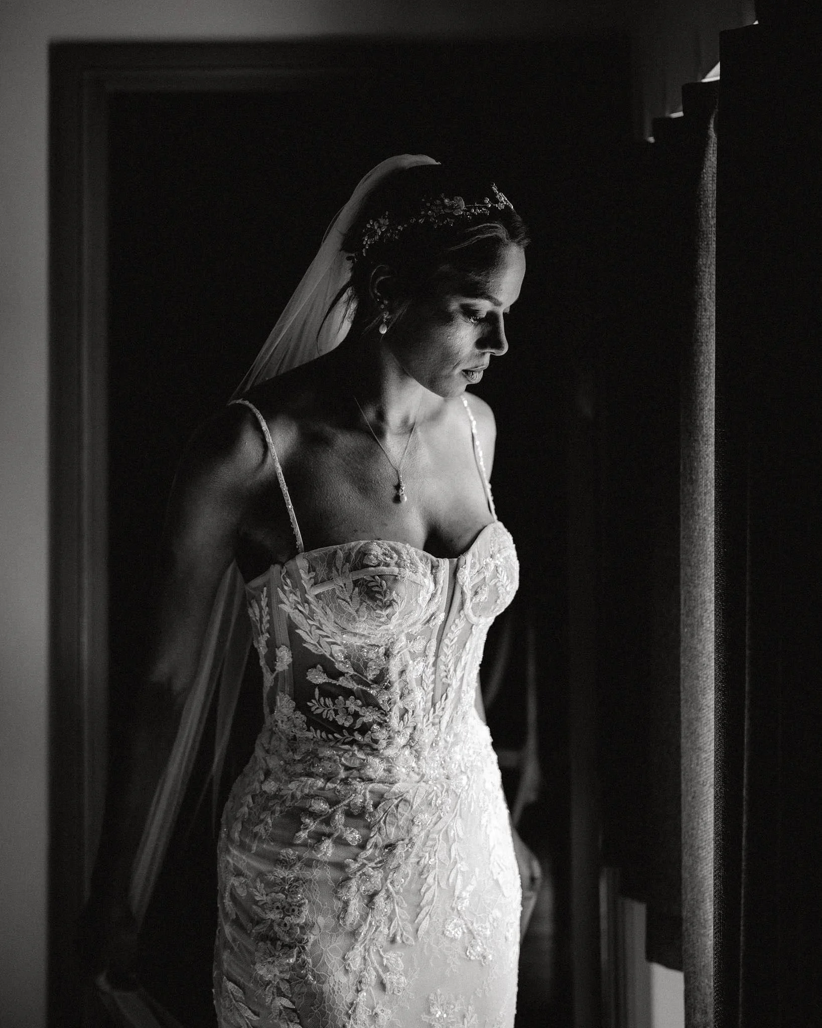 A bride in a lace wedding gown, with a veil and a headpiece, looking down thoughtfully in black-and-white photograph.