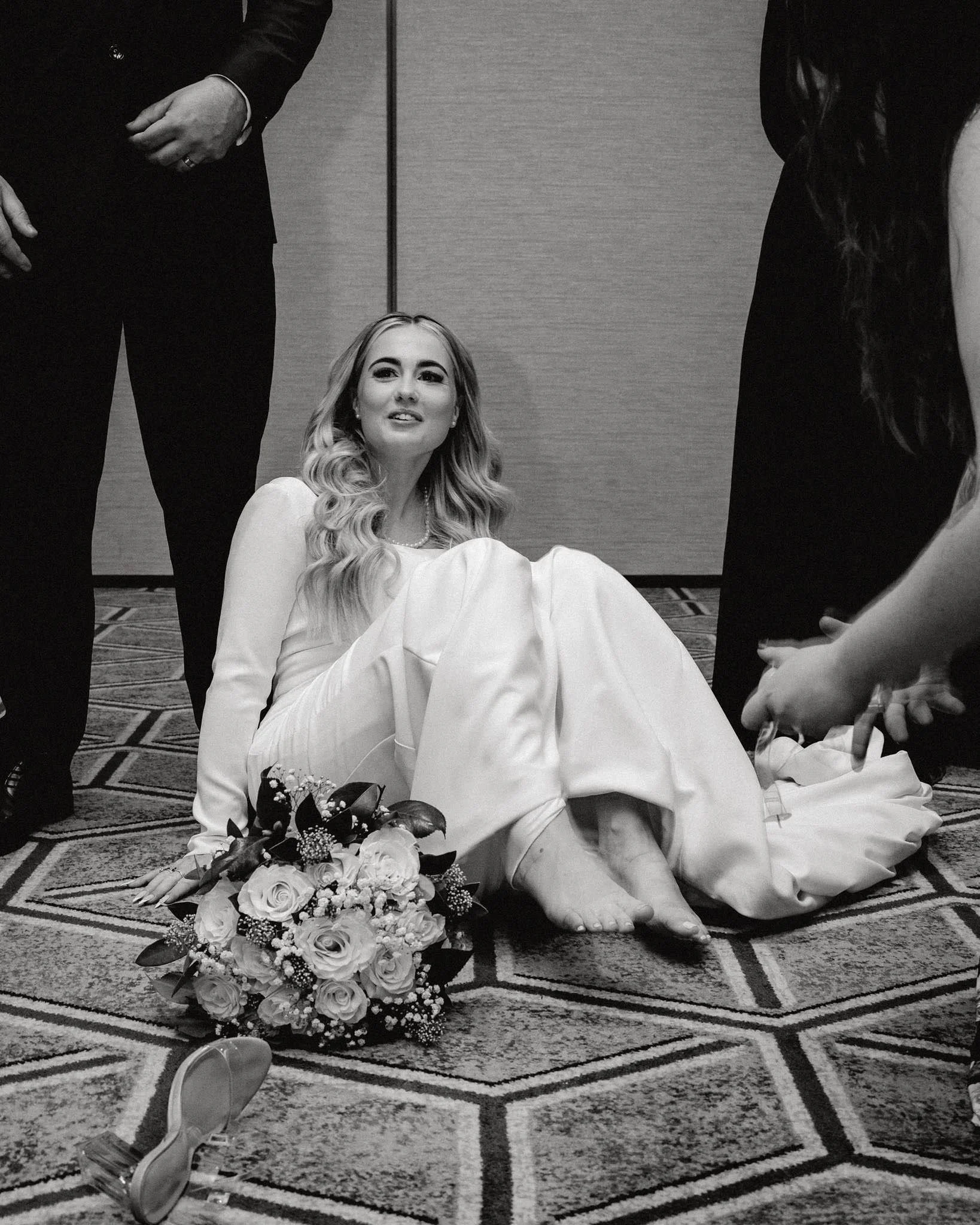 Woman in wedding dress sitting on the floor with bouquet of flowers, surrounded by people, in a formal setting.