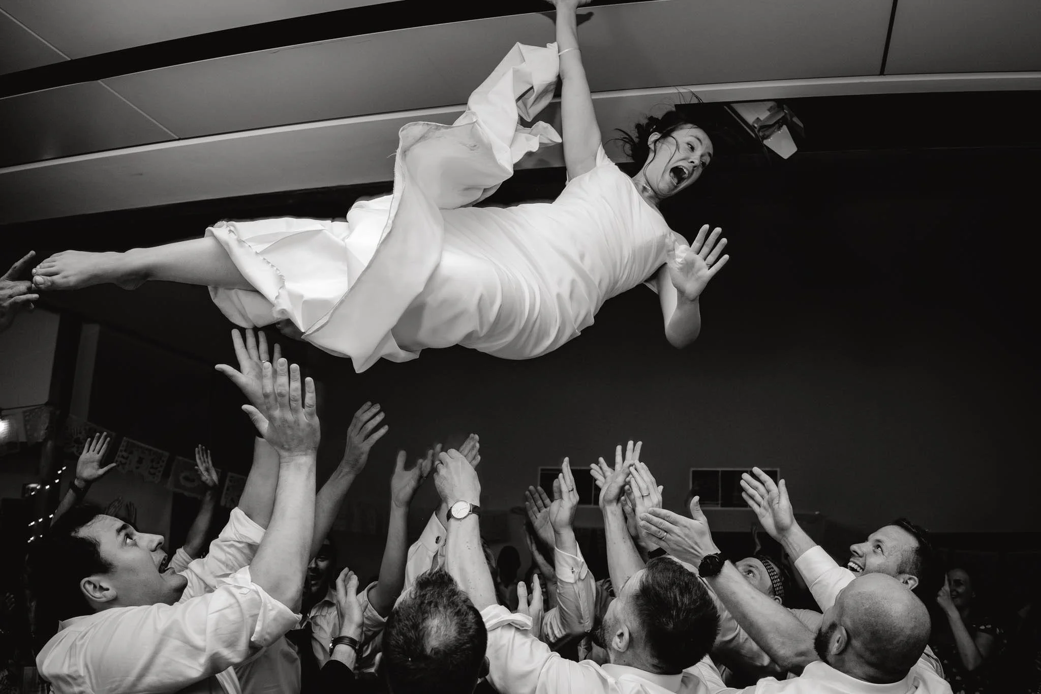 A woman in a wedding dress being lifted by a group of people at a celebration, with her mouth open in excitement.