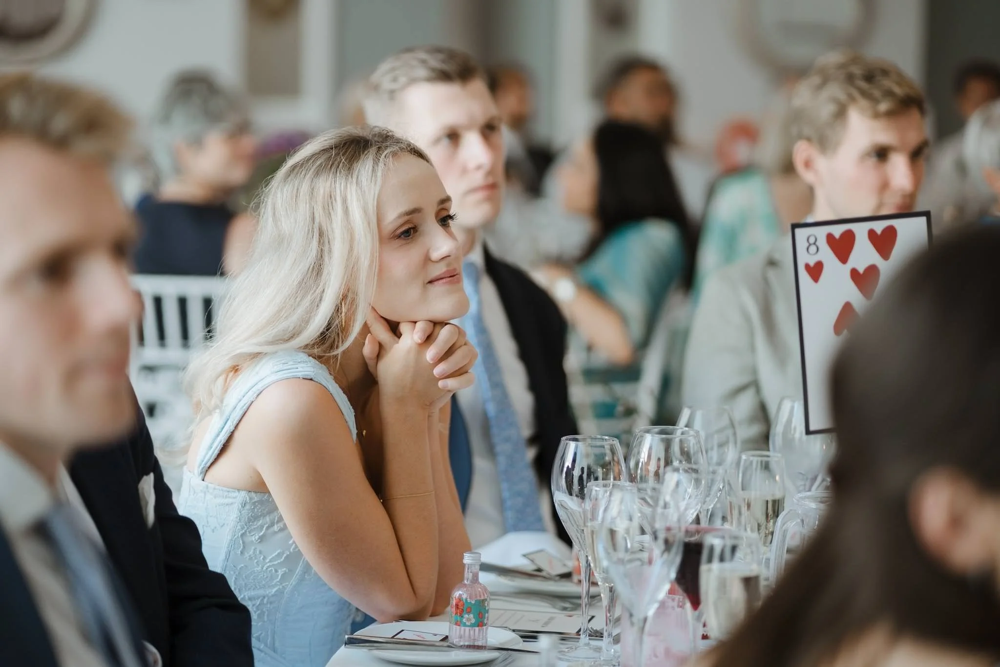 A young woman with blonde hair sitting at a formal event, attentively listening with her hands clasped under her chin. She is wearing a light blue dress and is surrounded by other elegantly dressed people at a table with wine glasses and a playing ca