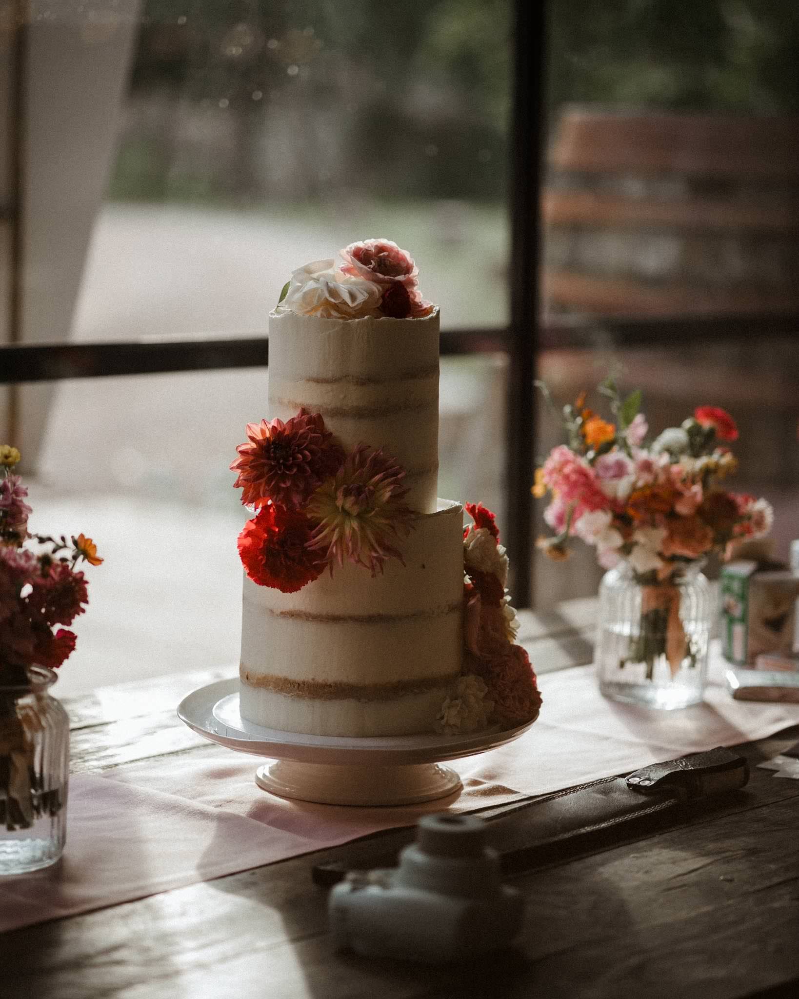 A three-tiered wedding cake decorated with pink and white flowers on a table with vases of flowers, in a rustic setting with natural light.