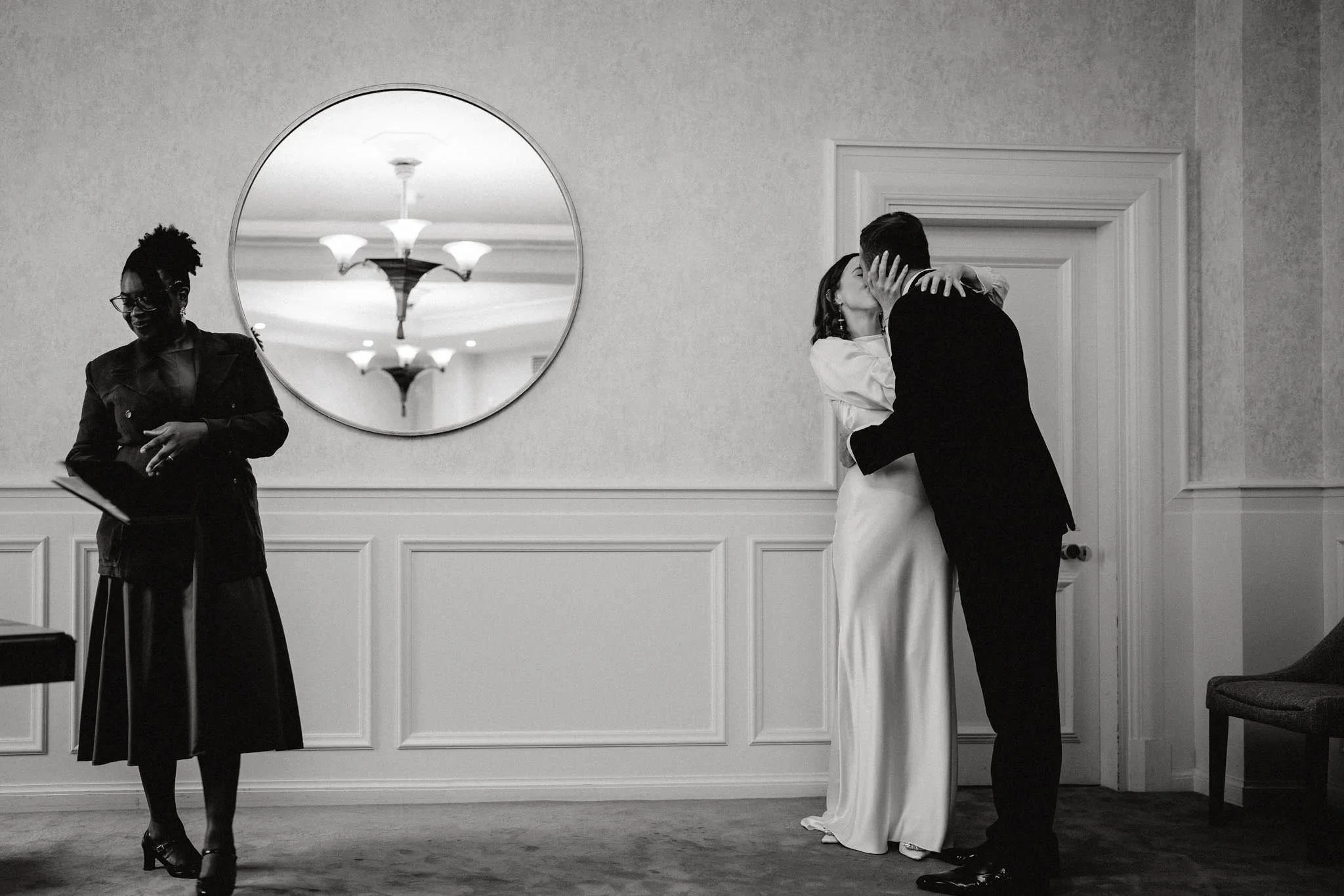 A couple kissing in a room with wainscoting, a round mirror on the wall, and a woman standing nearby holding a book or folder.
