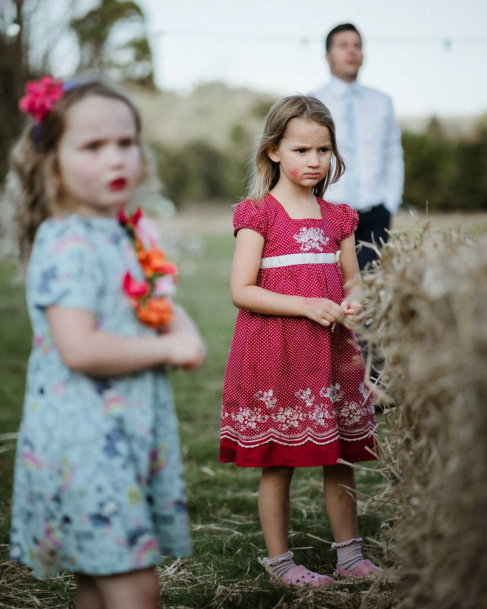 Three children, two girls and one man, stand outdoors near a haystack, with the girl in the center looking upset, and the girl on the left blurred in the foreground.