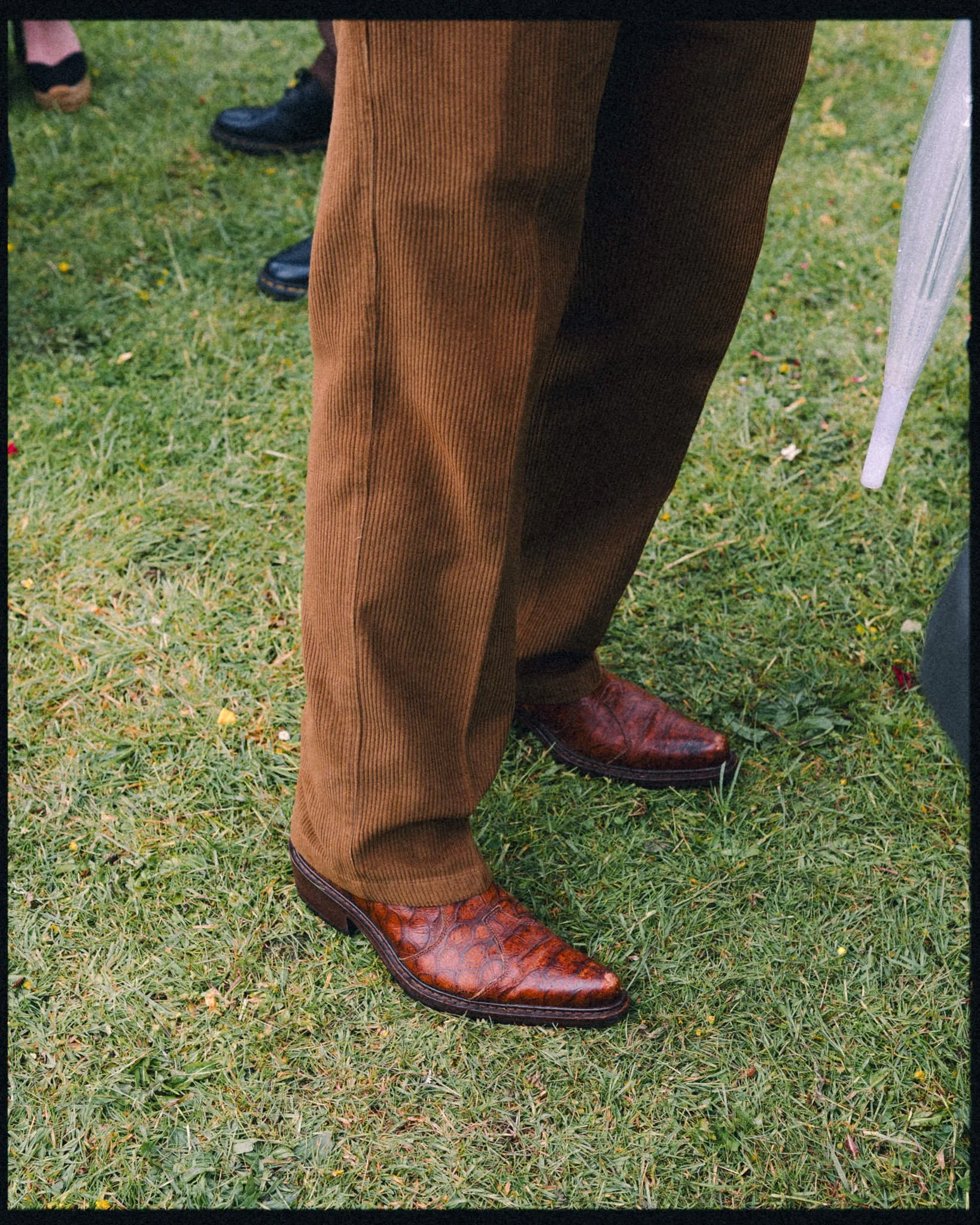 wedding guest in brown cowboy boots and brown trousers standing on the grass, shot from the thigh down.