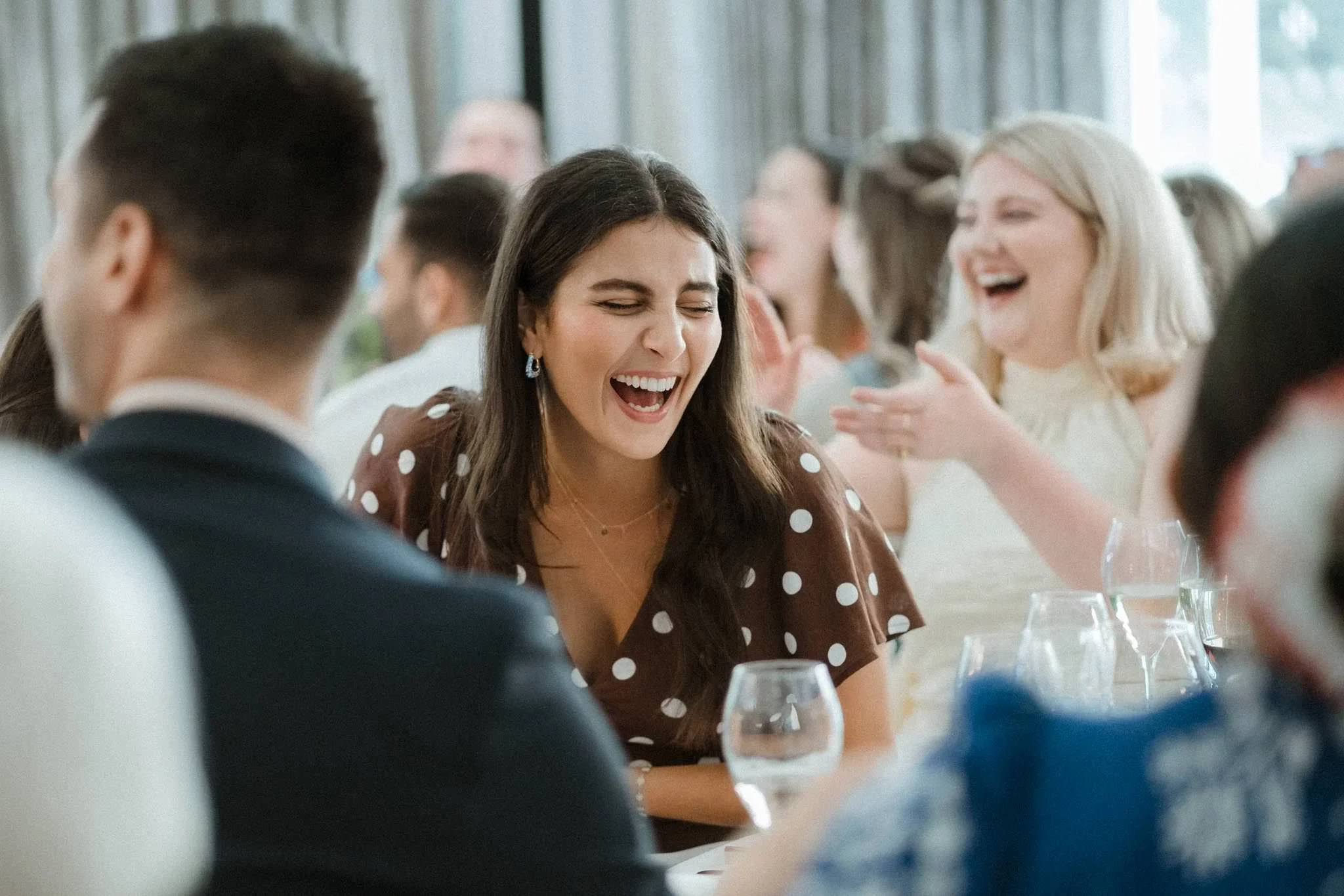 A group of people laughing and smiling at a social gathering or meal, seated at a table with wine glasses and dinnerware.