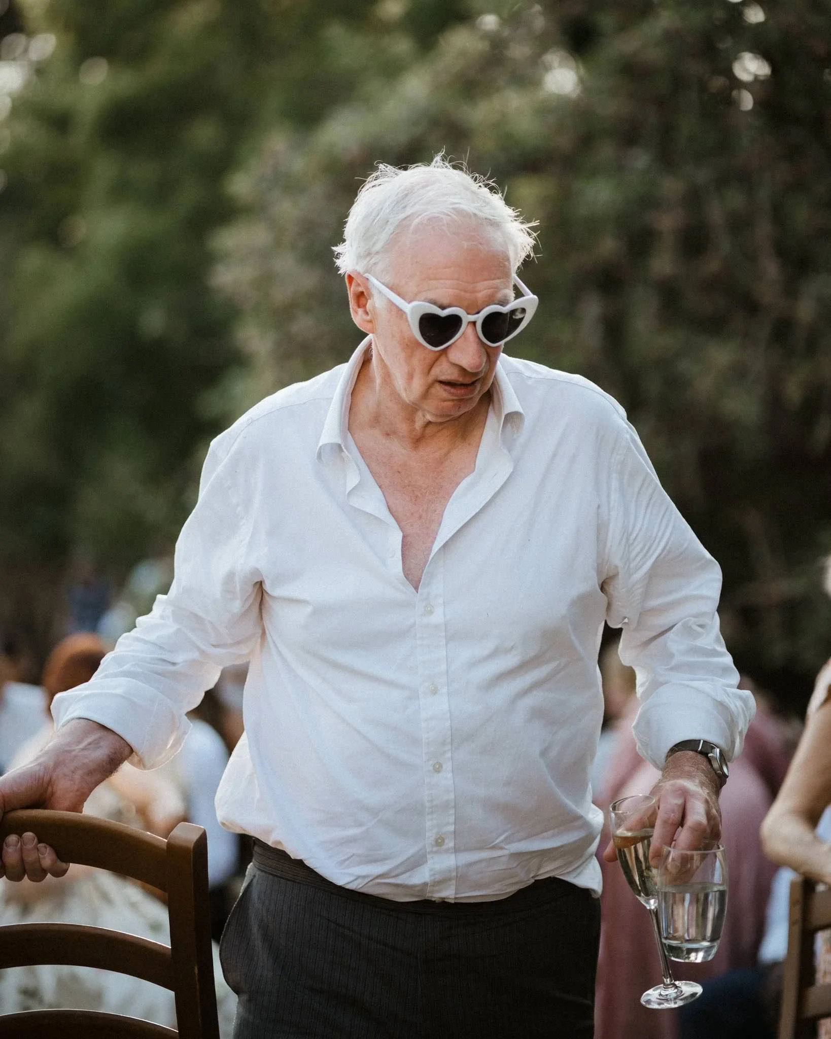 An elderly man with white hair and sunglasses with heart-shaped lenses, wearing a white shirt and dark pants, holding a glass of champagne and a glass of water at an outdoor event.