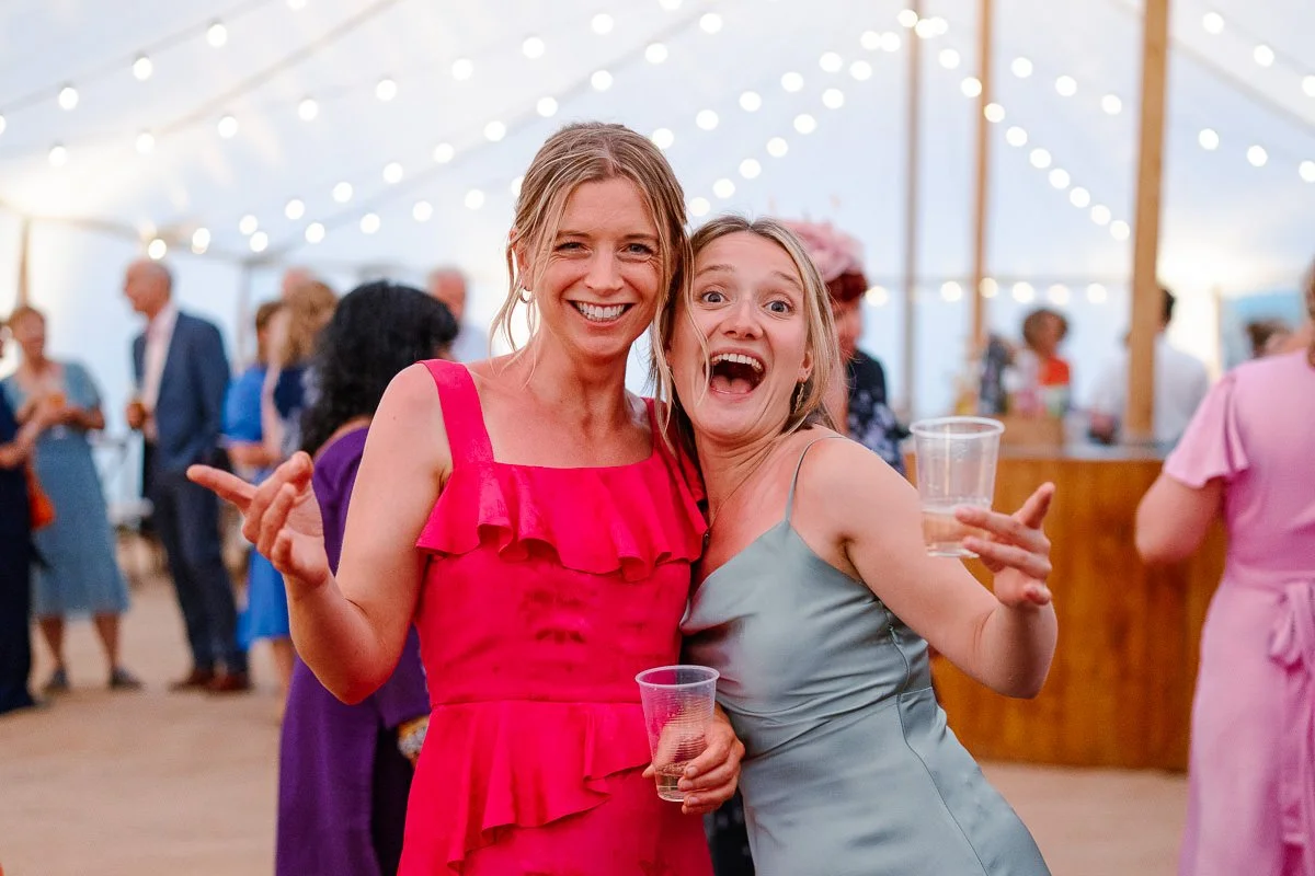 Two young guests in bright dresses laugh together at a marquee wedding, soft colour bokeh in the background