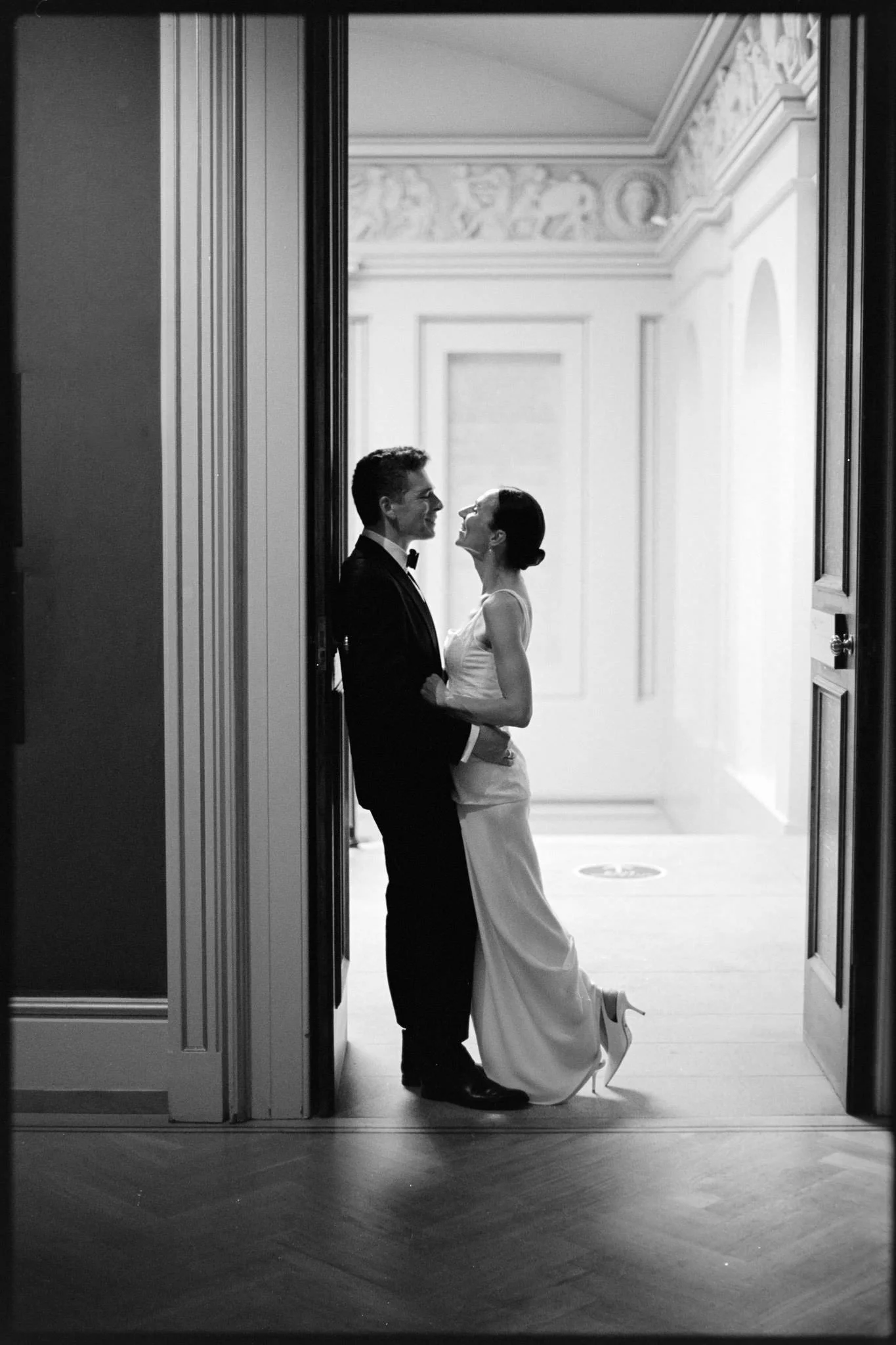 Bride and groom in a doorway at the ashmolean museum in Oxford, she lifts a leg before they embrace,  in black and white on film