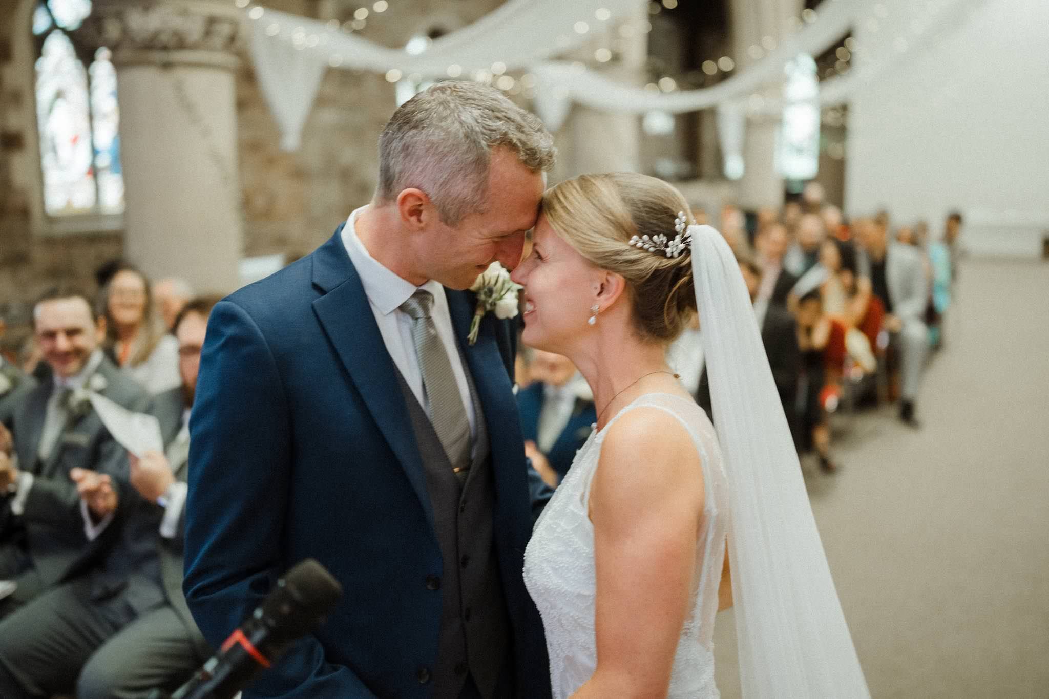 A bride and groom lean their foreheads together during their wedding ceremony inside a rustic stone church, surrounded by seated guests.
