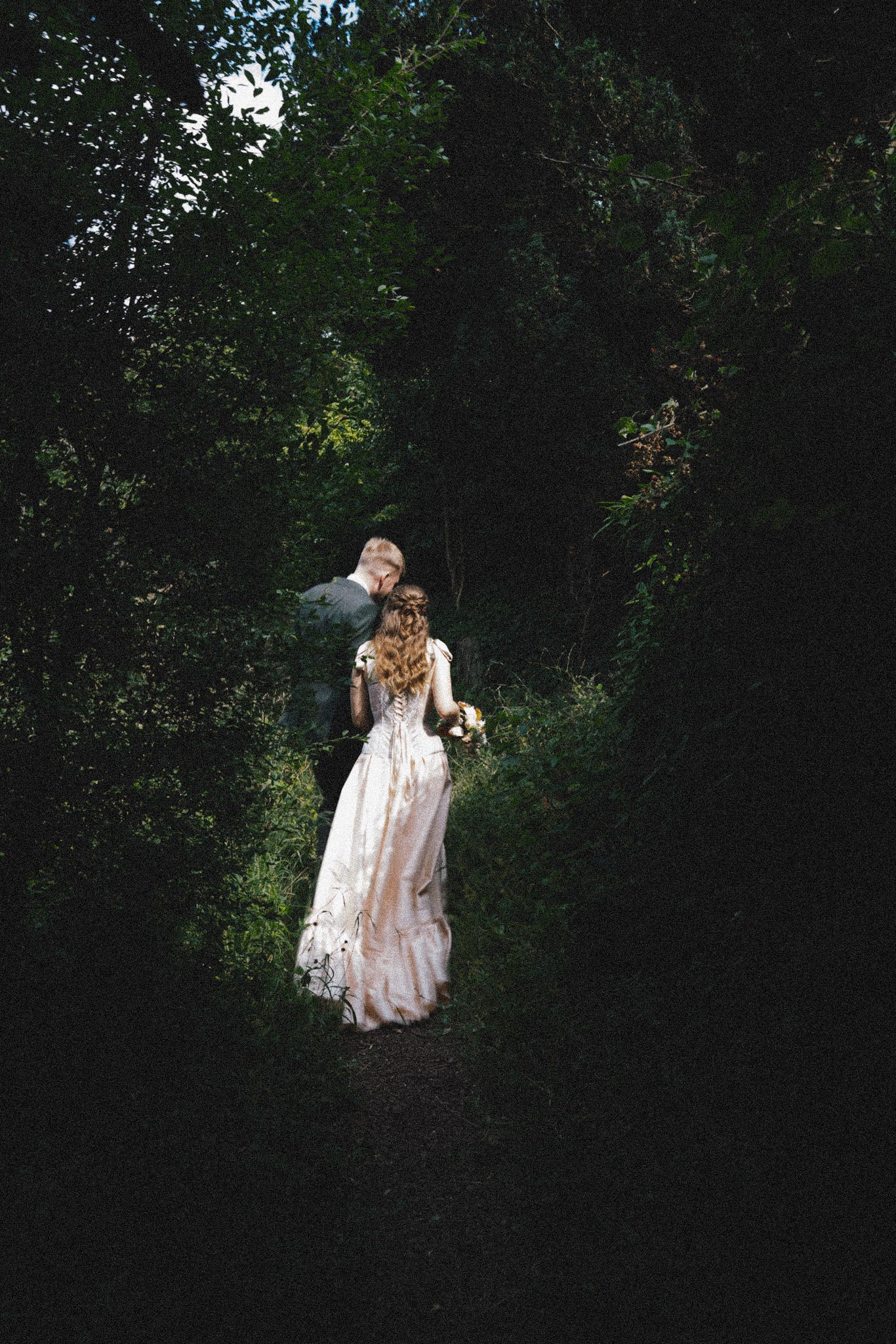 Couple disappear into the grounds at Arnos vale cemetery, they are lit by sunlight amongst dark trees as they walk away from the camera
