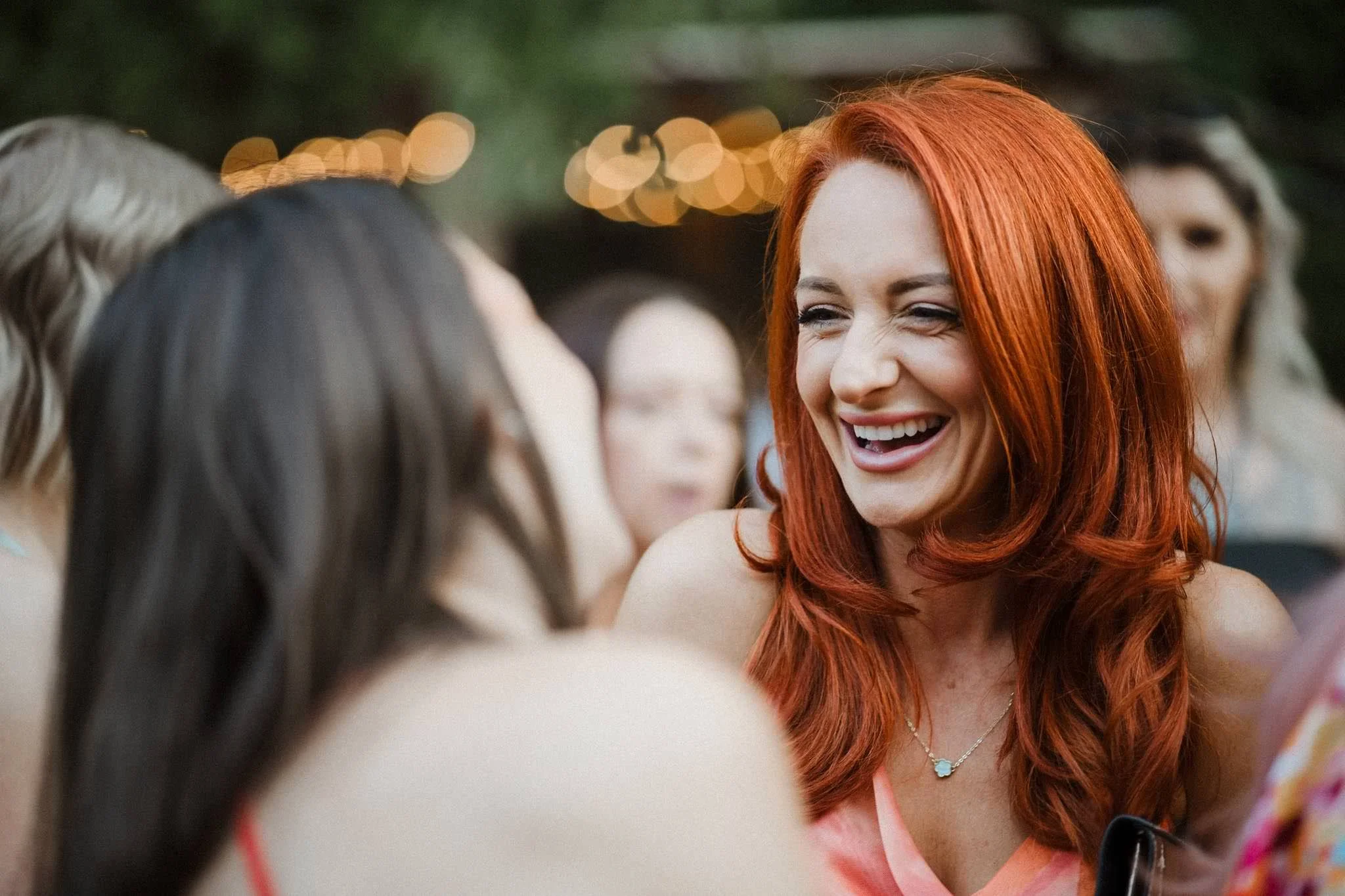A woman with long red hair smiling and talking to a group of women at an outdoor event in the evening with bokeh lights in the background.