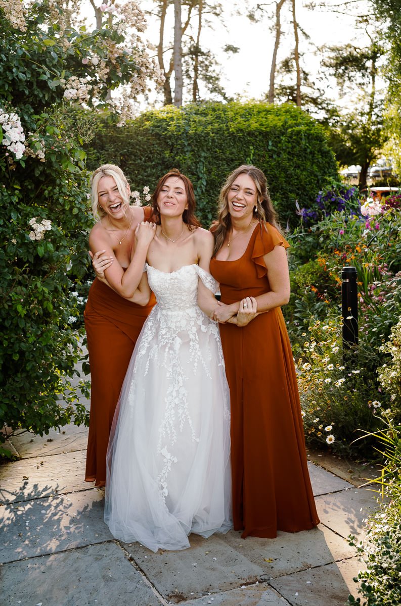 Bride shares a joyful laugh with friends surrounded by flowers at Kingscote Barn, captured in vibrant colour