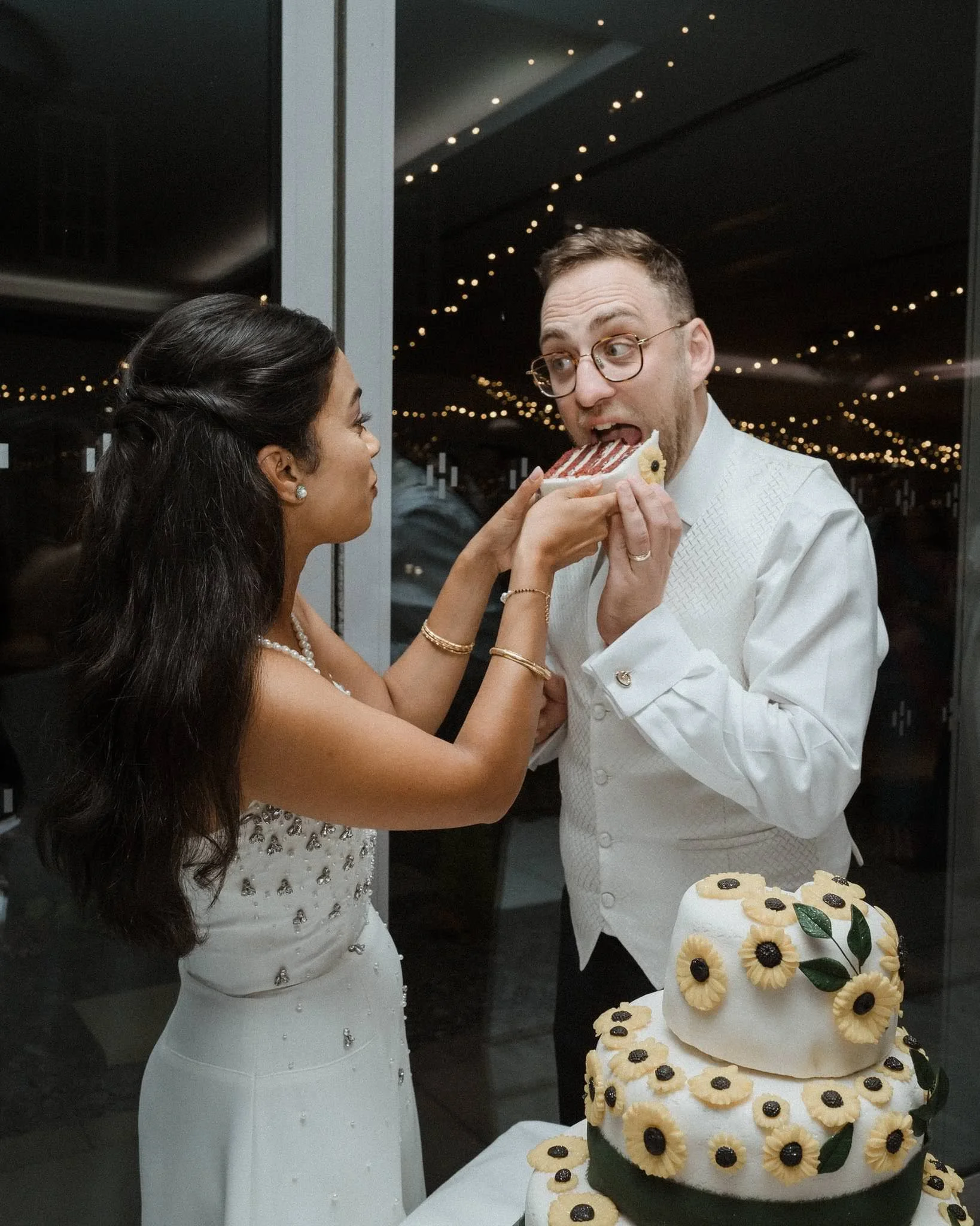 A bride and groom at their wedding reception, with the bride holding a slice of cake near the groom's mouth. The cake is decorated with sunflower and leaf designs.