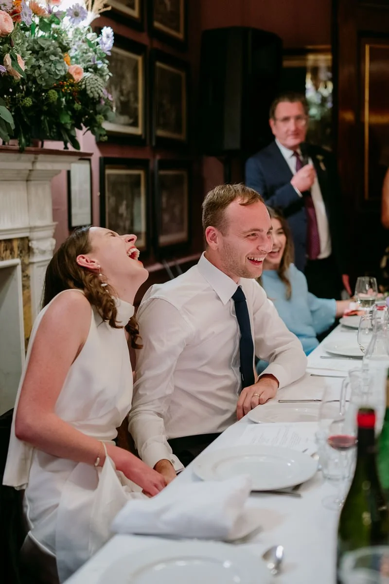 Bride bursts into laughter during her father's speech at the dinner table, Brunswick House, London — candid colour wedding photography