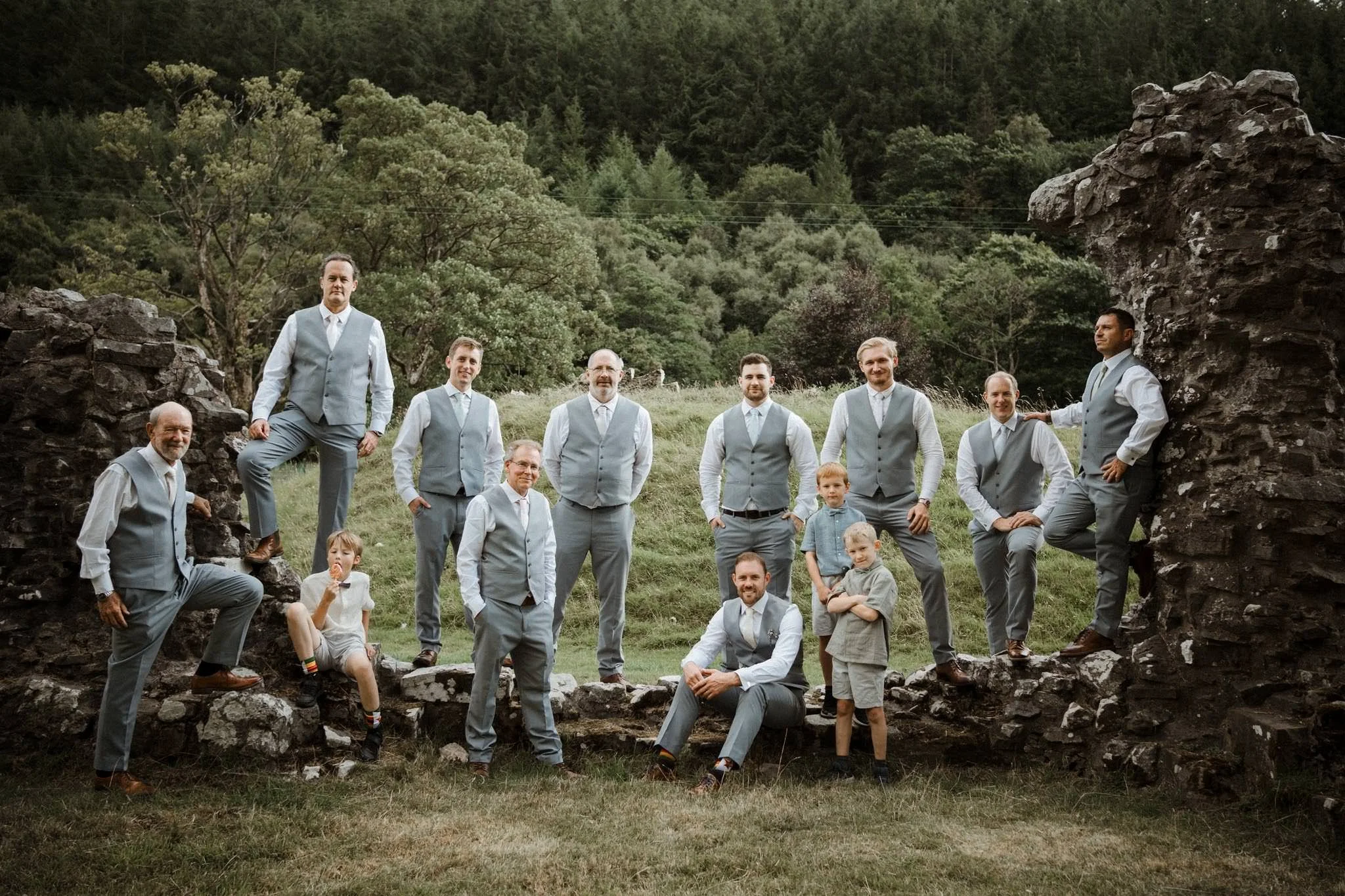 A group of men and a boy in gray suits and vests posing outdoors on a grassy area with stone ruins and green trees in the background.