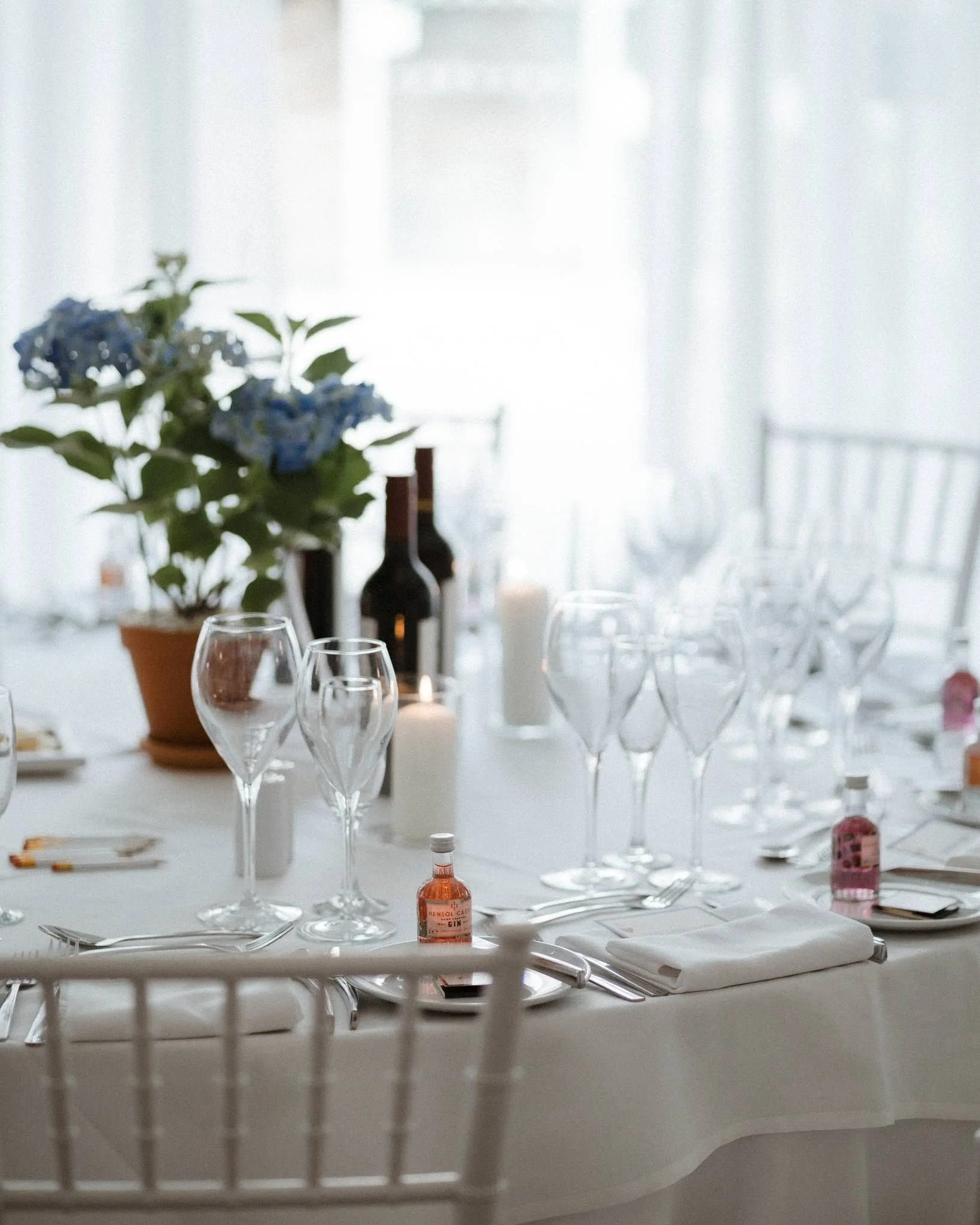 Elegant table set with white tablecloth, empty wine glasses, candles, a potted plant with blue flowers, and decorative bottles, in a bright room with sheer curtains.