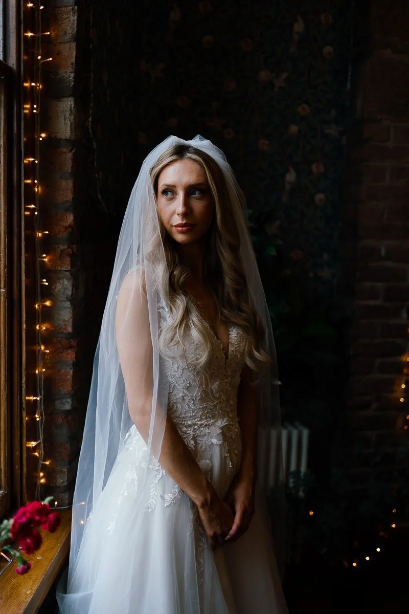 Bride poses by a window at the Radnor Rooms, Bristol, showing off the delicate lace detailing on her dress in soft colour light