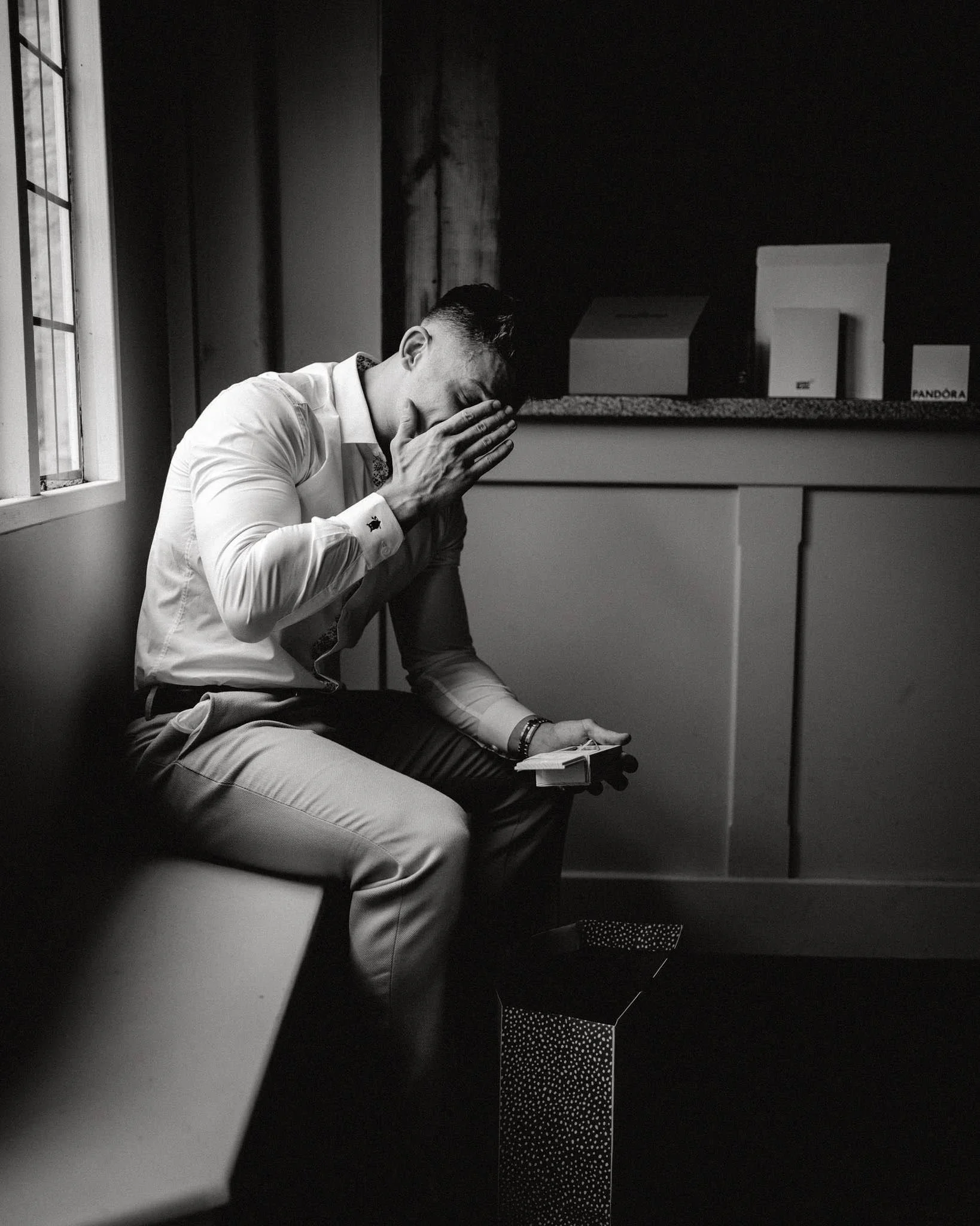 A man sitting on a bench near a window, covering his face with one hand and holding a small box in the other, appearing distressed.