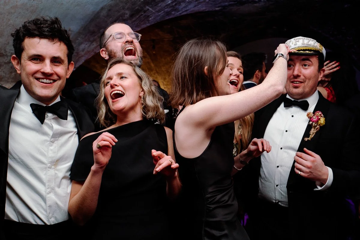 Guests dance and laugh in a Tux Fizz sailor hat, men in black tie and women in black — high-energy colour dancefloor photography