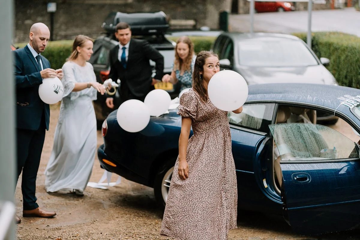 Wedding guest blows up a white balloon beside a classic Jaguar at Woodlands Church, a playful candid moment in colour