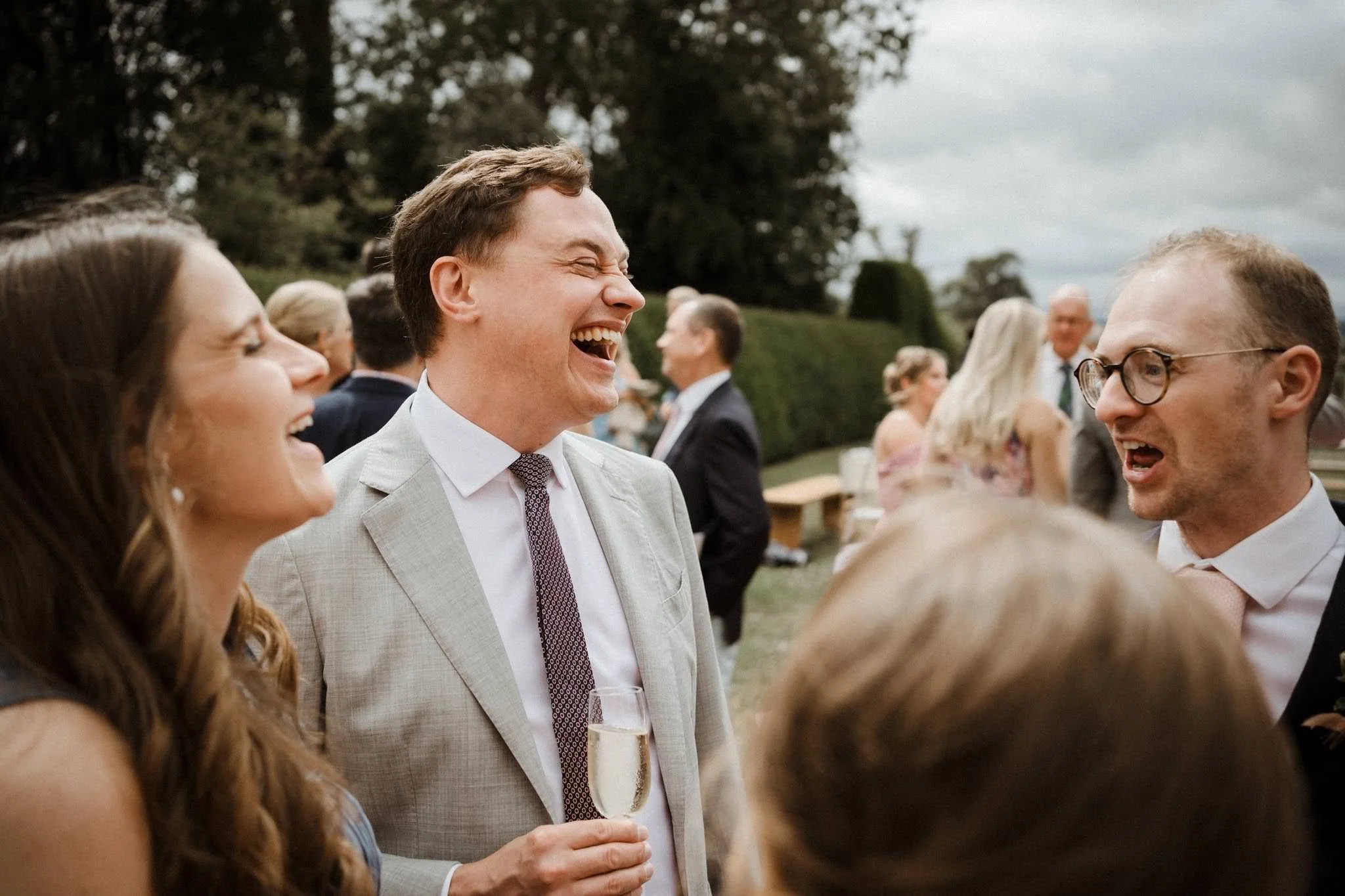 Group of people at an outdoor wedding reception, laughing and talking, some holding champagne glasses, with a lush green background and cloudy sky.