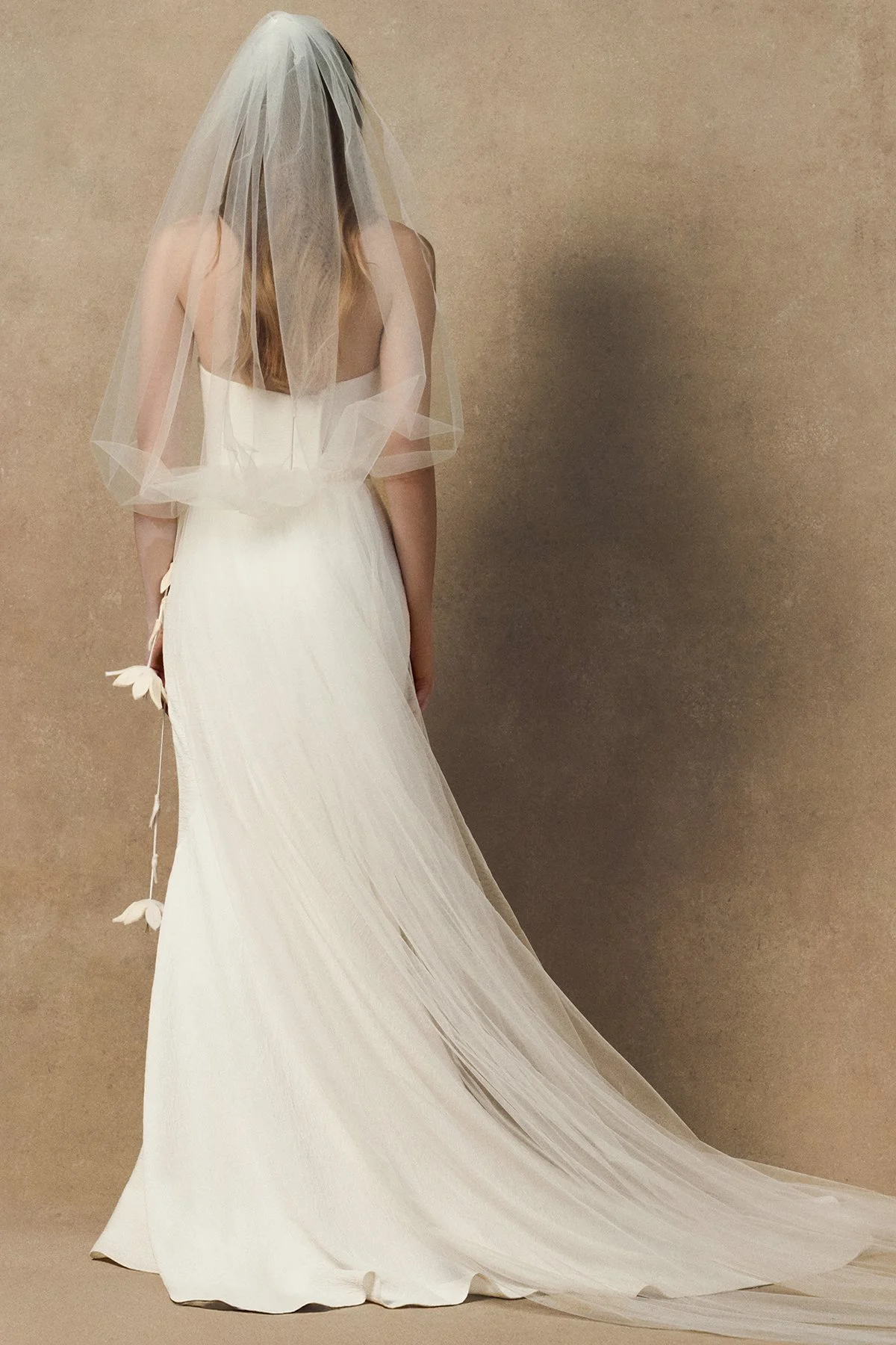 Back view of the Phyllis Veil, a floor-length sheer tulle bridal veil with a soft gather at the crown, falling over the shoulders. tucked into waistband and worn with nicole dress
