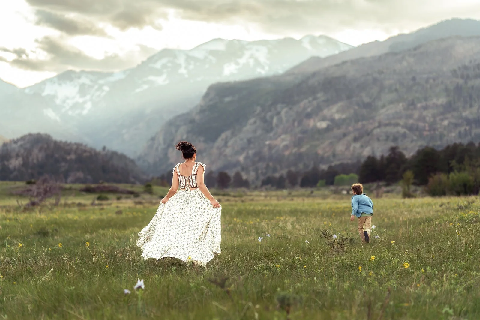 RMNP Photographer | Mother walks away from the camera into a big meadown with sun running. Mountains are in front of them at Moraine Valley in RMNP.