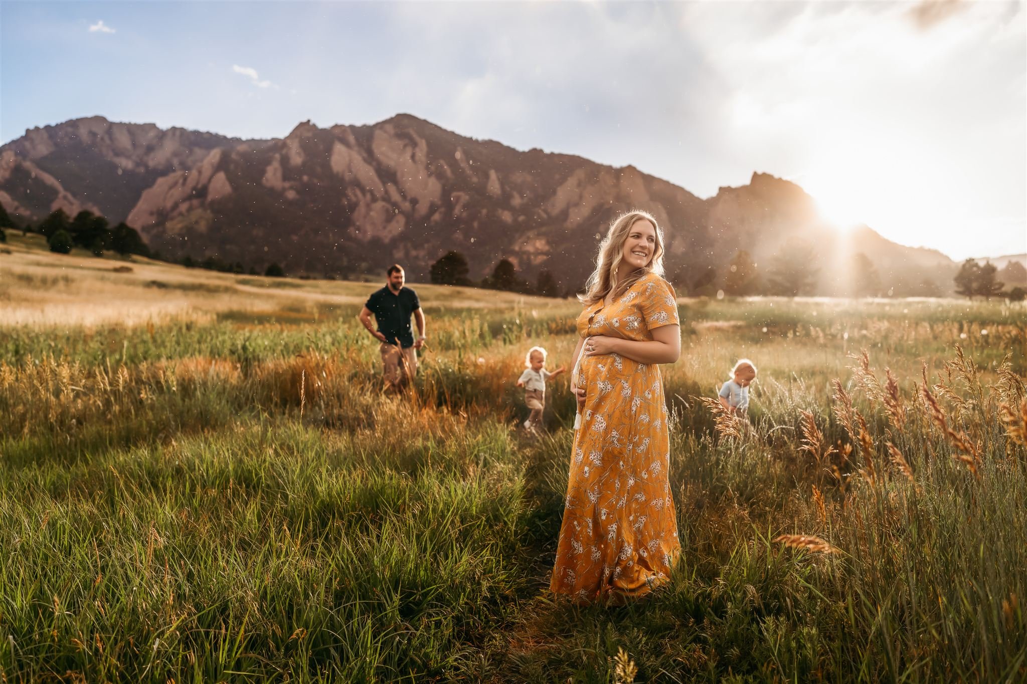 Denver Family Photographer | Pregnant Mother stands in yellow dress holding her belly while the kids and husband run through behind her. It's sunny and raining.