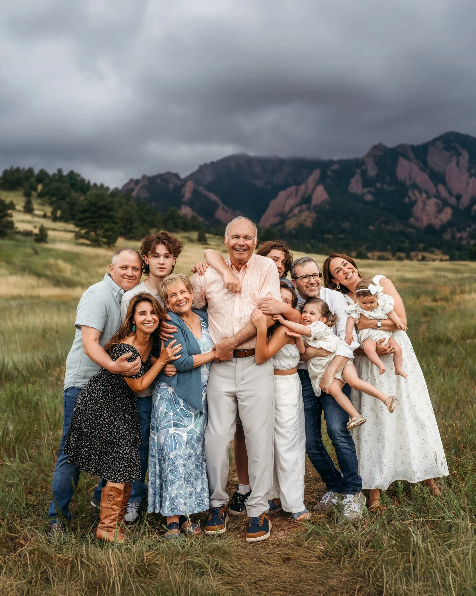 Extended Family Photography in Boulder | An extended family all together, looking at the camera and smiling and laughing on green grass in front of the flatirons in Boulder. everyone is huddled around the grandfather.